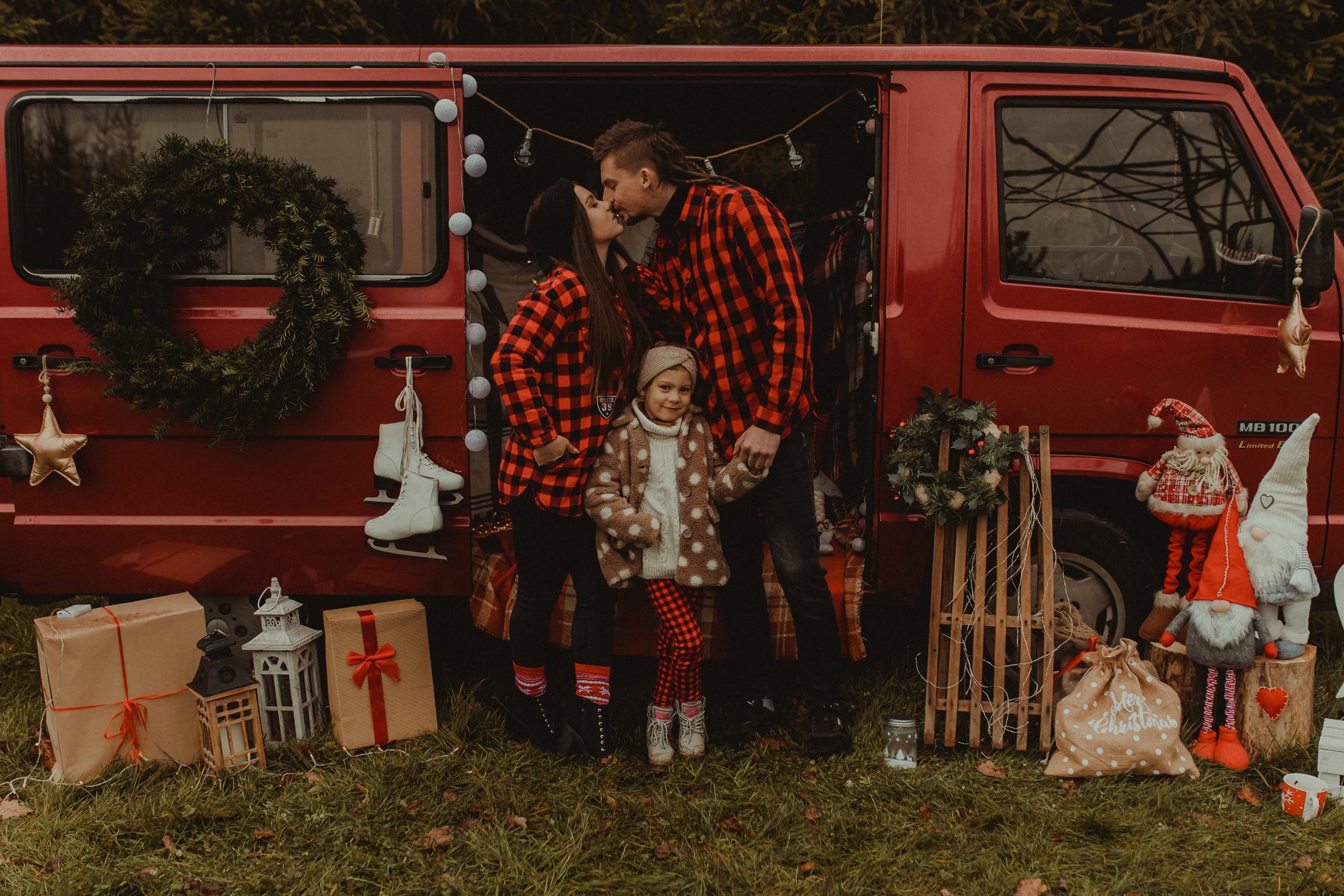 Rodzice całują się przed świątecznym busem. Przed nimi stoi dziewczynka w kurtce w grochy
