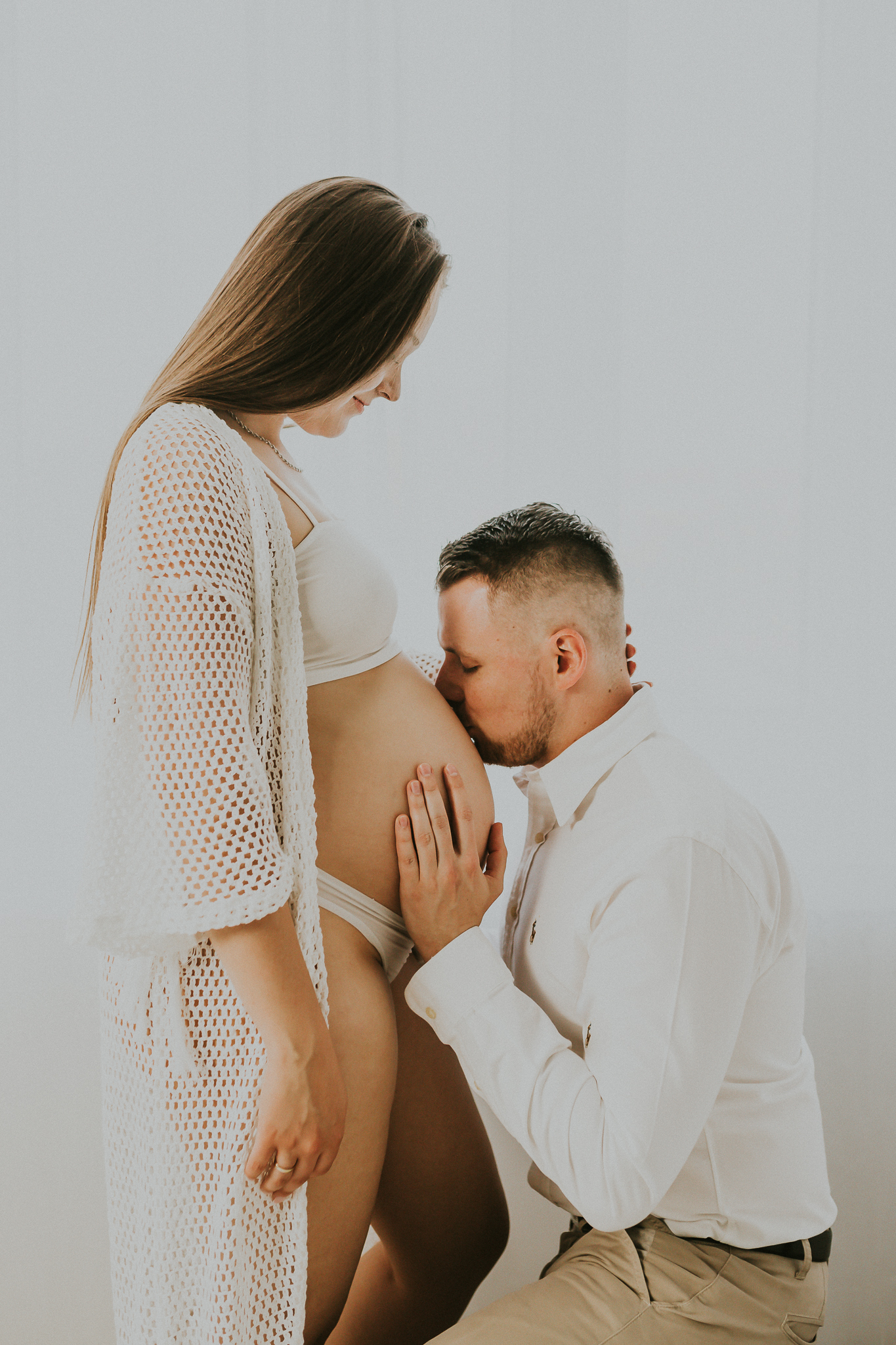 Mężczyzna całuje brzuch ciążowy kobiety. Ubrani na biało