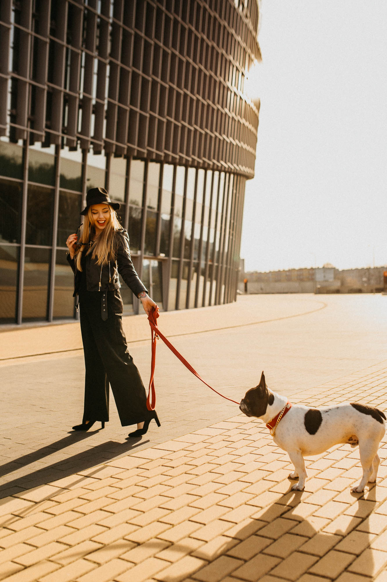 Blondynka ubrana na czarno, trzyma psa na czerwonej smyczy w tle nowoczesna architektura