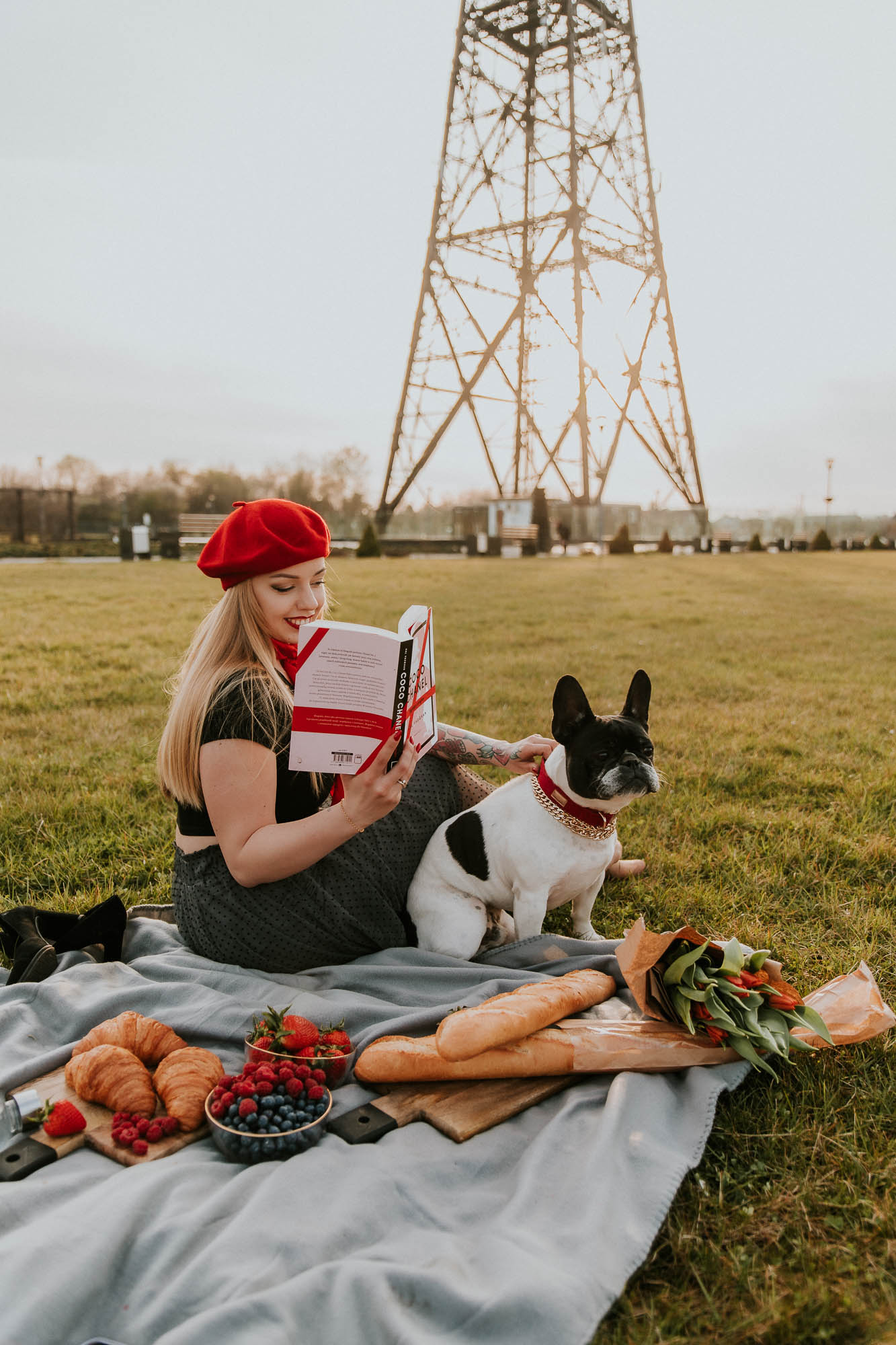Dziewczyna w czerwonym berecie na kocu czyta książkę, a obok siedzi pies