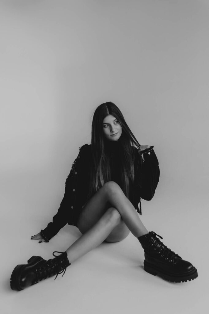 Brunetka siedząca przy ścianie z założonymi nogami w czarnej stylizacji, ma gołe nogi, Fusialka, fotografia czarno- biała