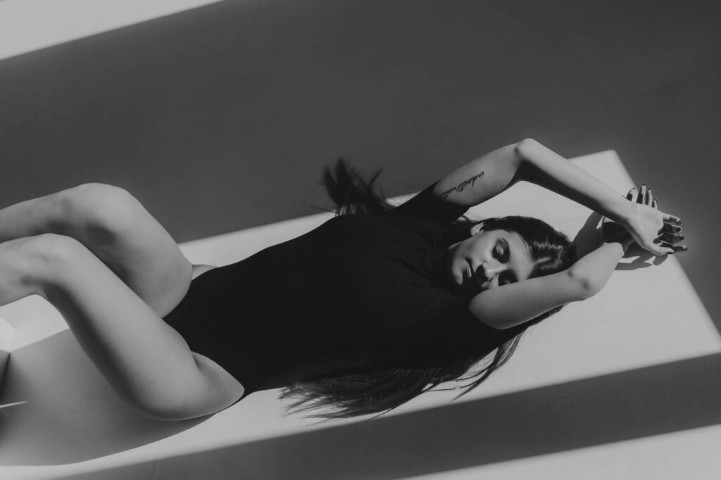 Kobieta leżąca na podłodze w czarnym body, ręce ma skrzyżowane za głową. Fusialka. Zdjęcie czarno- białe.