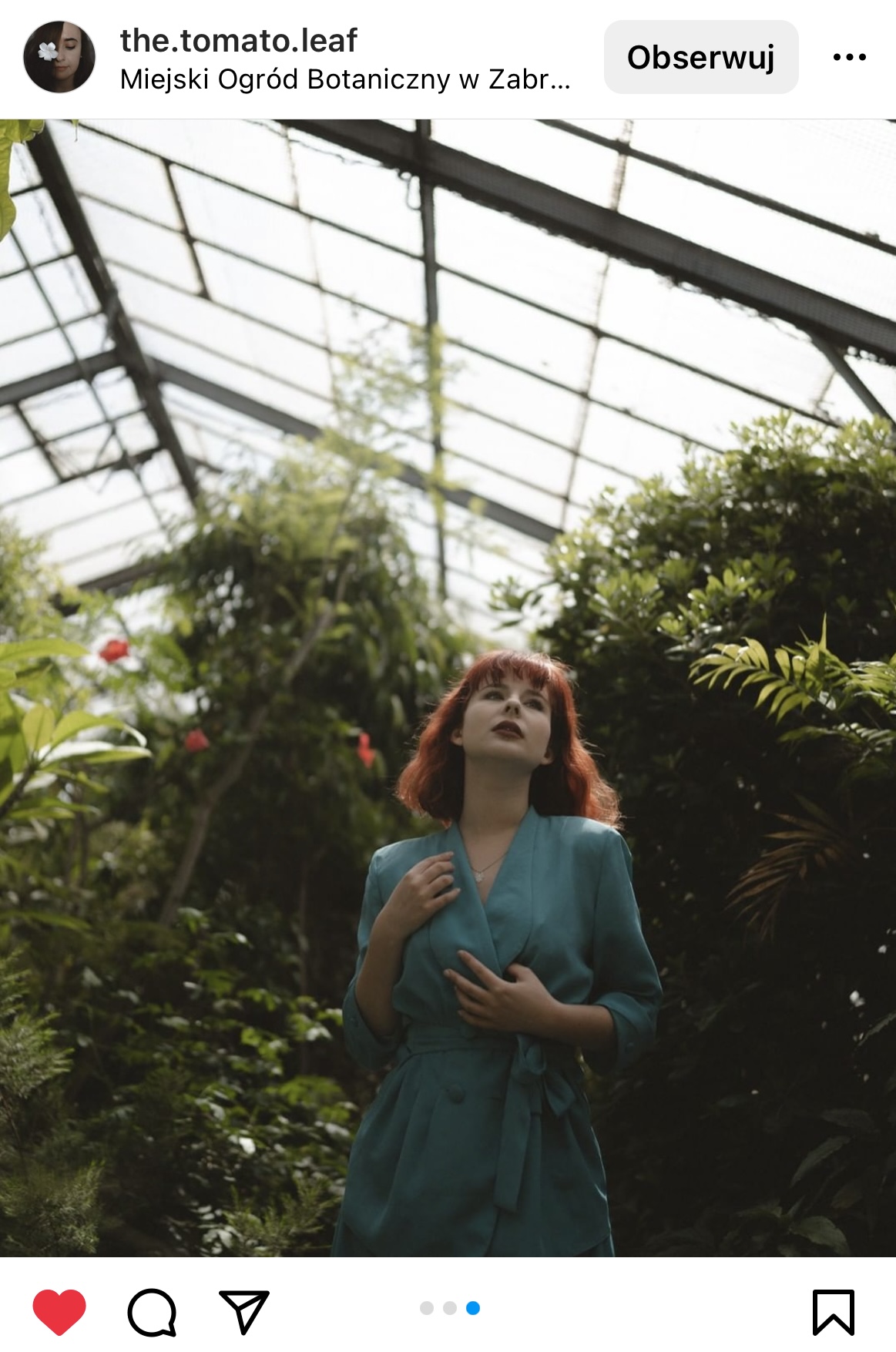 Kobieta w seledynowej sukience i rudych włosach stoi w szklarni pośród roślin.