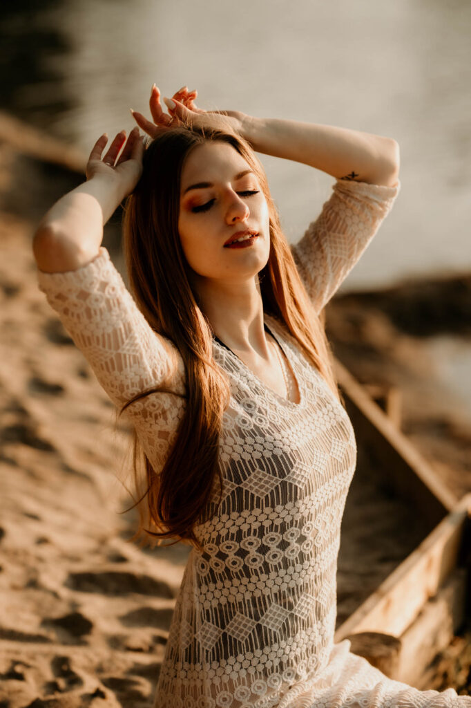 młoda kobieta trzyma ręce na głowie i uśmiecha się, ma zamknięte oczy, siedzi na brzegu jeziora