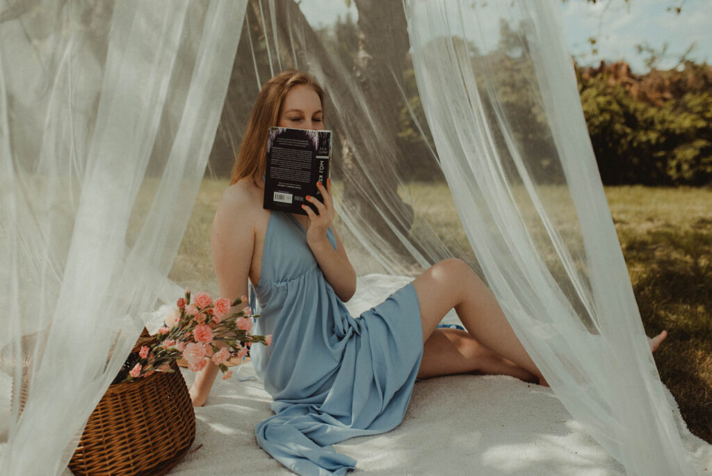 młoda kobieta w błękitnej sukience zakrywa twarz książką, obok kosz pełen kwiatów