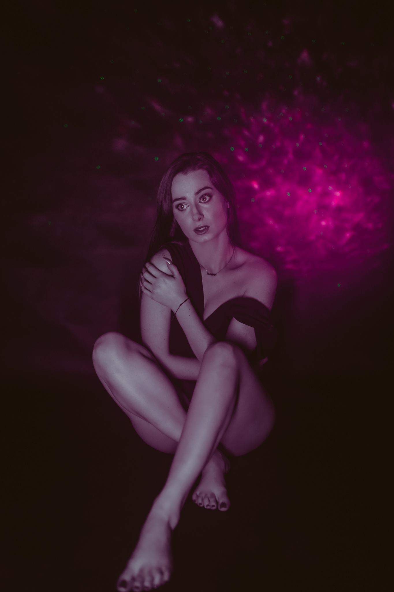 Kobieta na tle fioletowego koloru ściany. Minę ma zaciekawioną, siedzi z nogami skrzyżowanymi