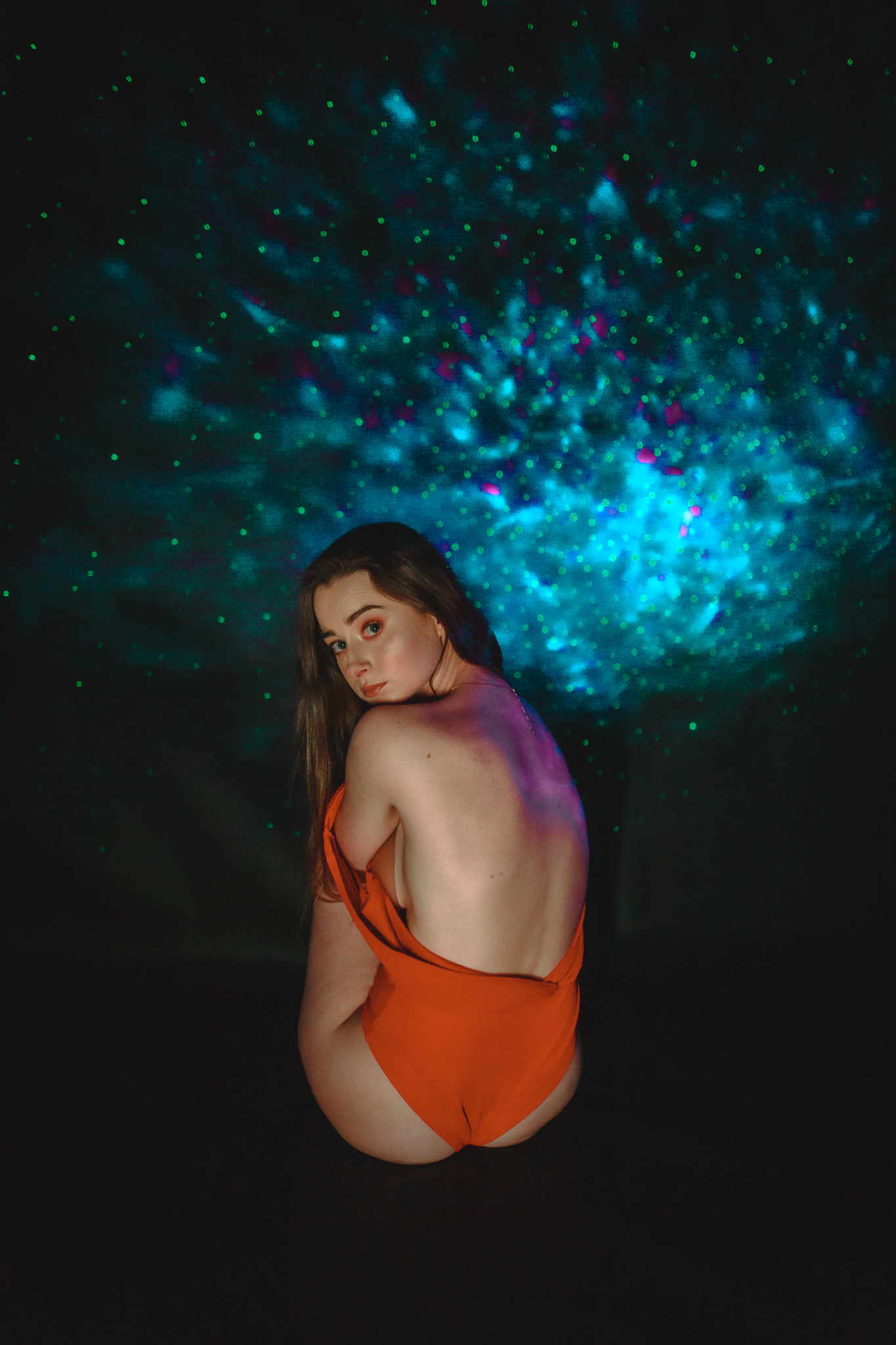 Młoda kobieta, która siedzi tyłem do obiektywu, głowę ma odwróconą do zdjęcia. Ramiączko opada jej na ciało