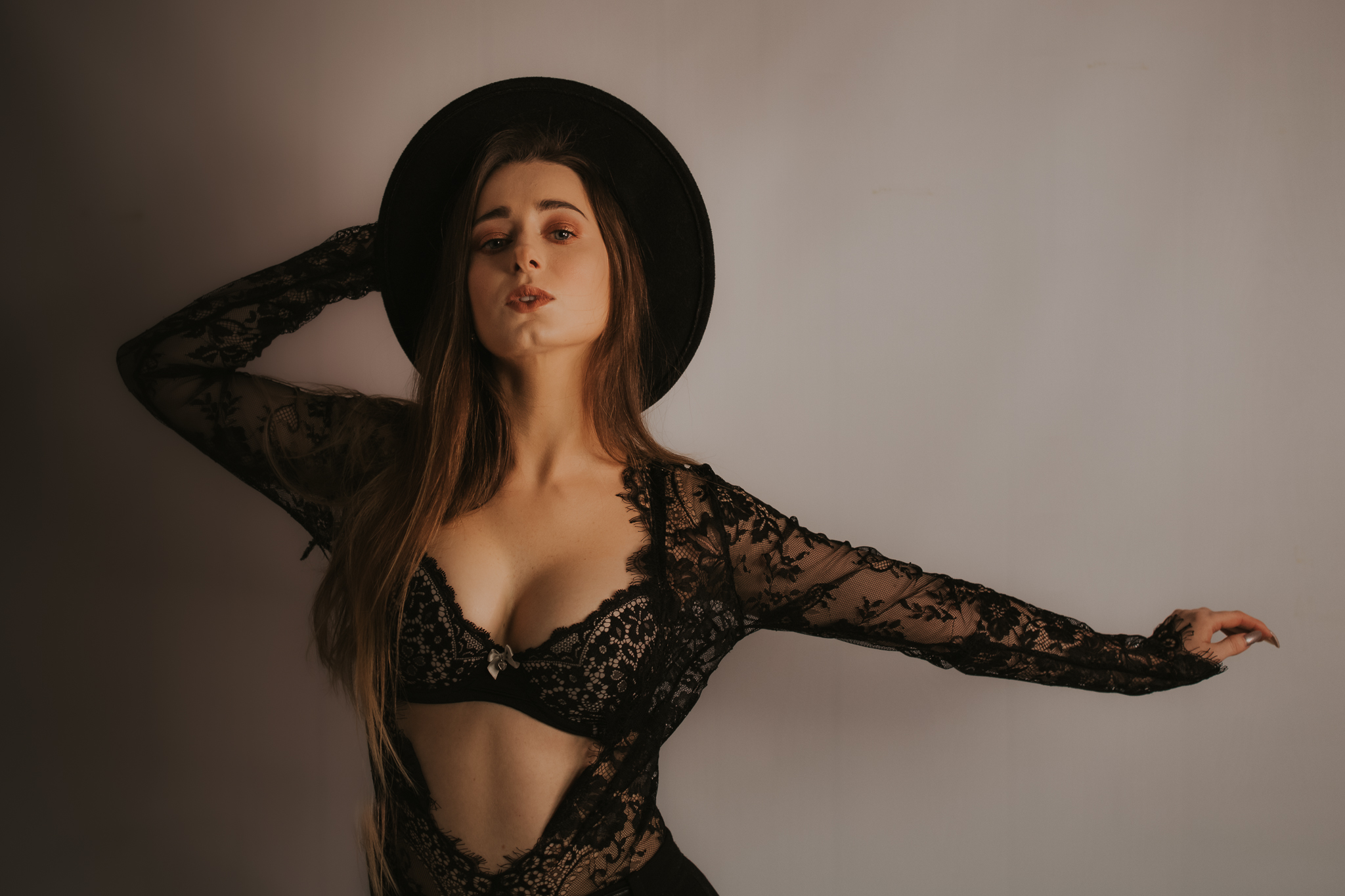 Kobieta w kapeluszu jedną rękę ma wyciągniętą, drugą trzyma kapelusz. Eksponuje piersi w pięknym, koronkowym staniku