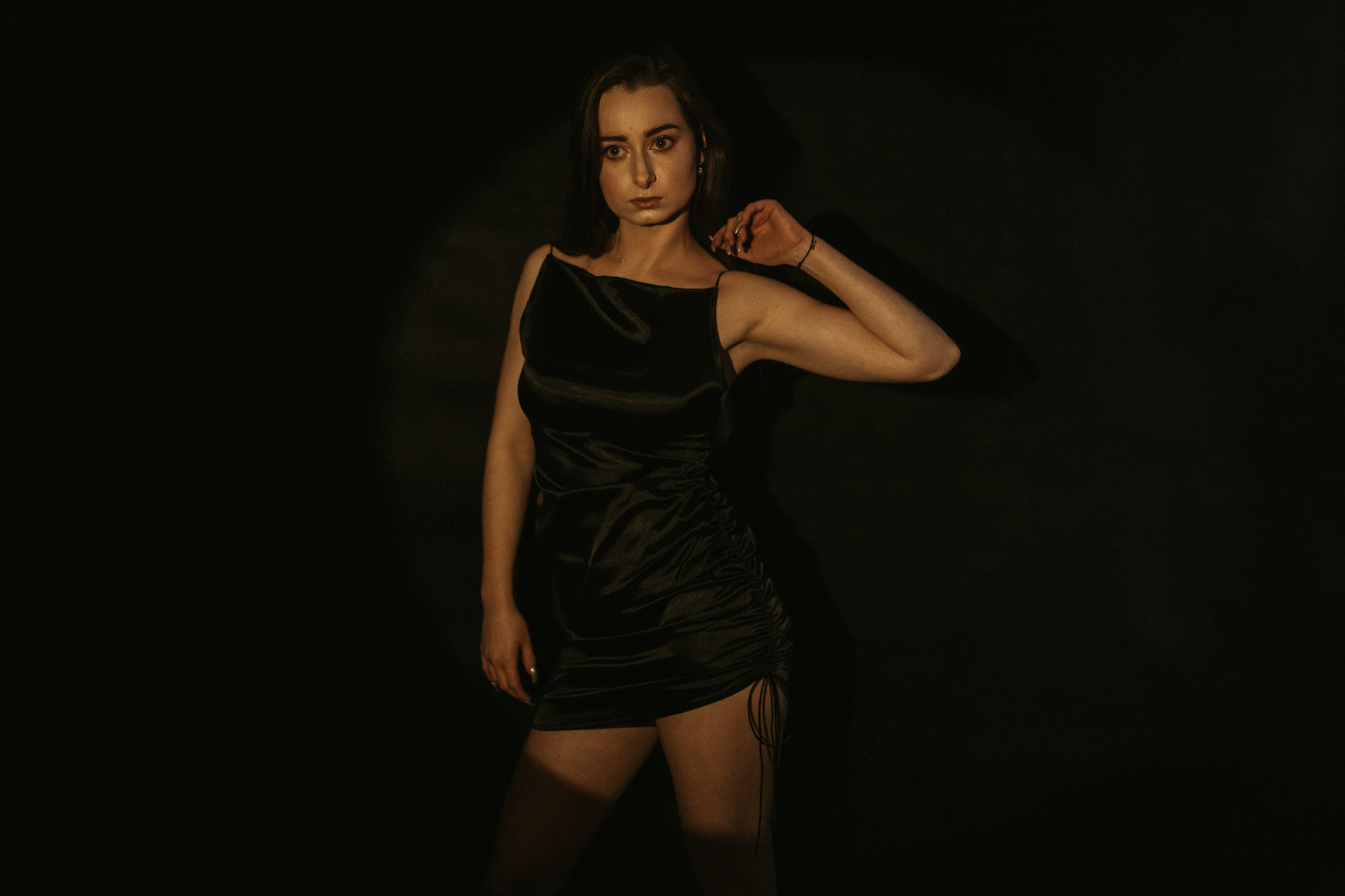 Kobieta stojąca na czarnym tle z ręką uniesioną ku głowie. Ma krótką, czarną spódniczkę