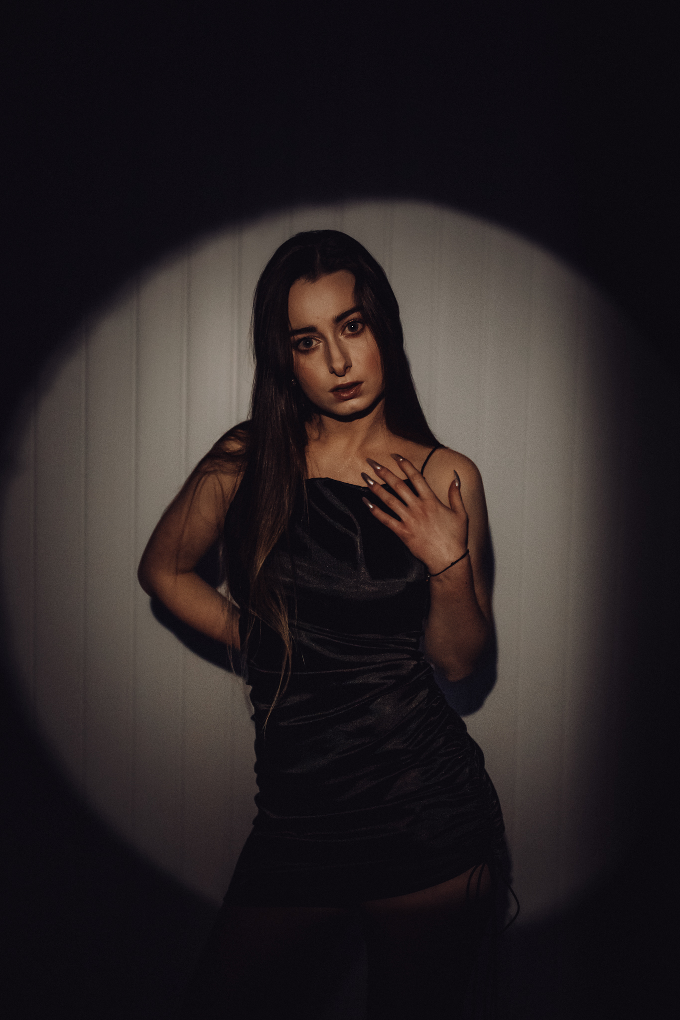 Brunetka w czarnej bluzeczce na ramiączkach stoi przy ścianie i patrzy w obiektyw. Jedną rękę ma na dekoldzie drugą za sobą