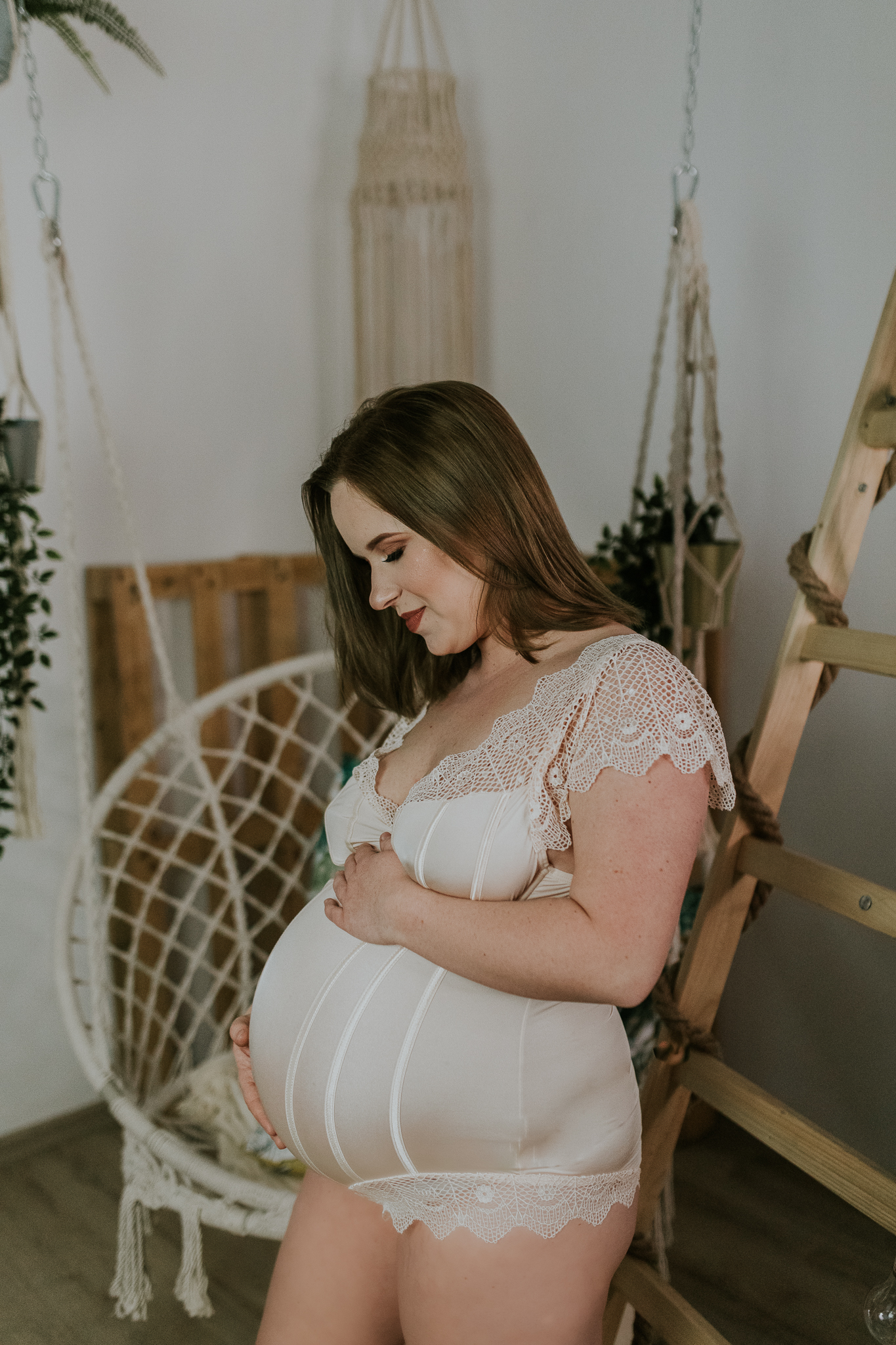 Kobieta w białym ciążowym body siedzi na huśtawce boho i trzyma się za ciążowy brzuch