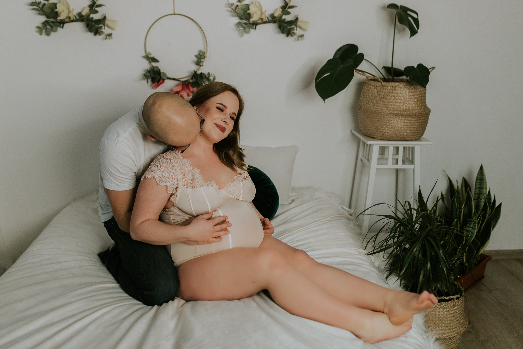 Para siedzi na łóżku. Mężczyzna całuje kobietę po szyi i obejmuje ją. Sesja ciążowa