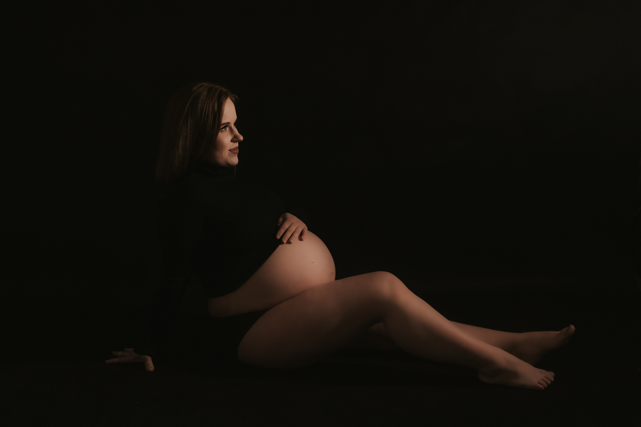 Kobieta siedzi na podłodze, patrzy przed siebie i trzyma się za brzuch. Sesja ciążowa