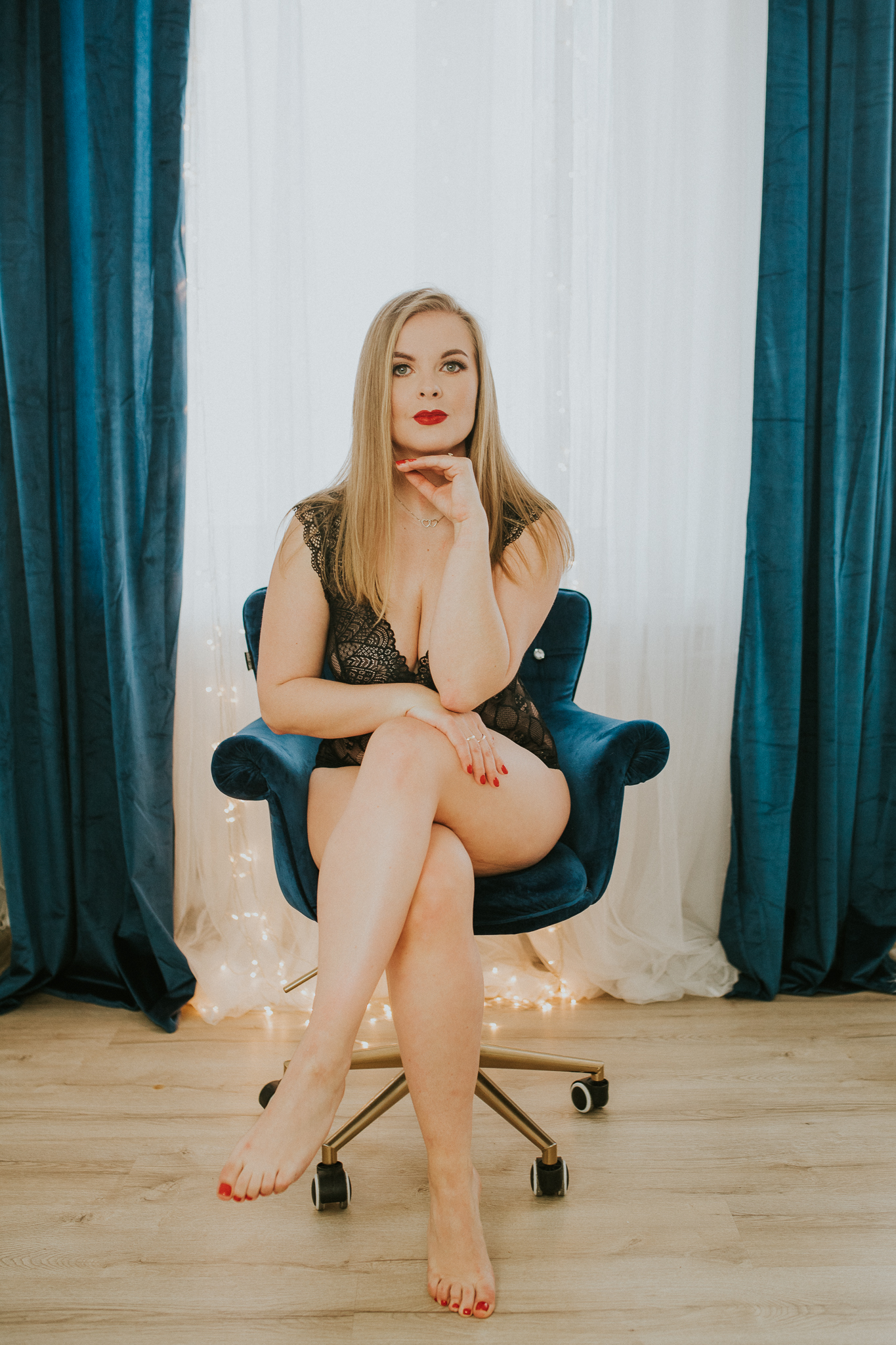 Kobieta siedząca na turkusowym fotelu w czarnym body z ręką pod brodą