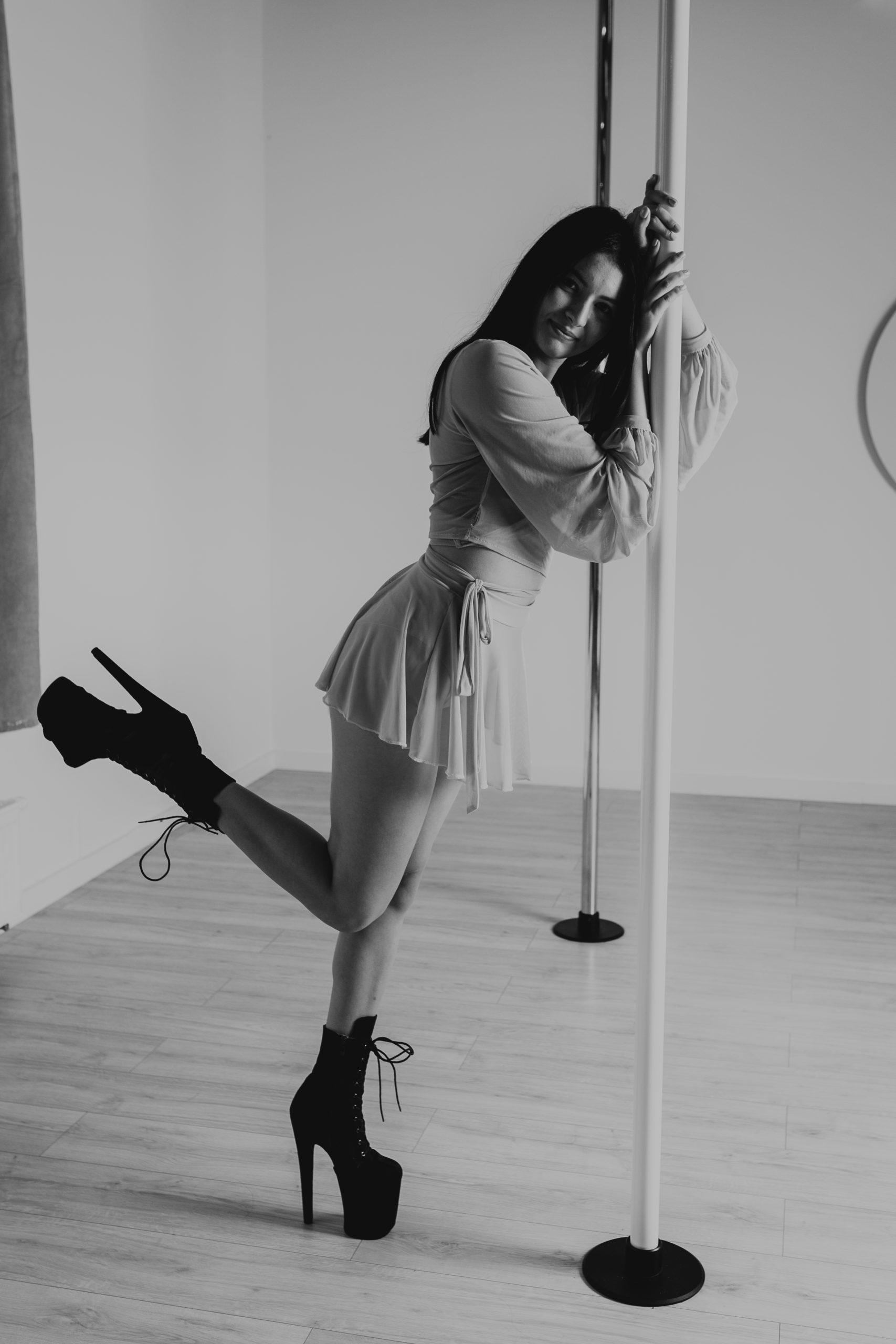 Dziewczyna w sukience i butach na wysokim obcasie trzyma się rury. Nogę ma uniesioną i zgiętą w kolanie