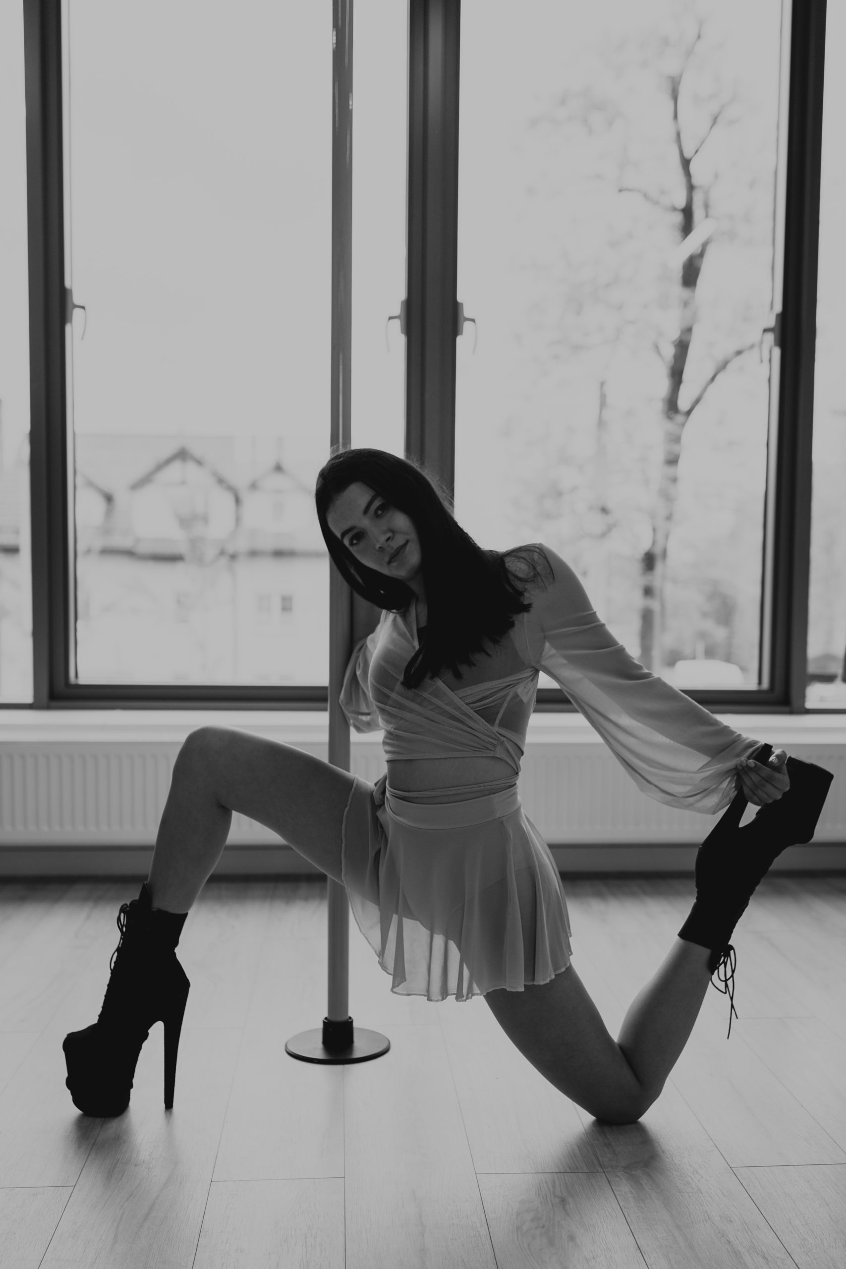 Piękna kobieta w sukience oparta jest o podłogę kolanem. Ma wysokie buty na obcasie i seksowną sukienkę