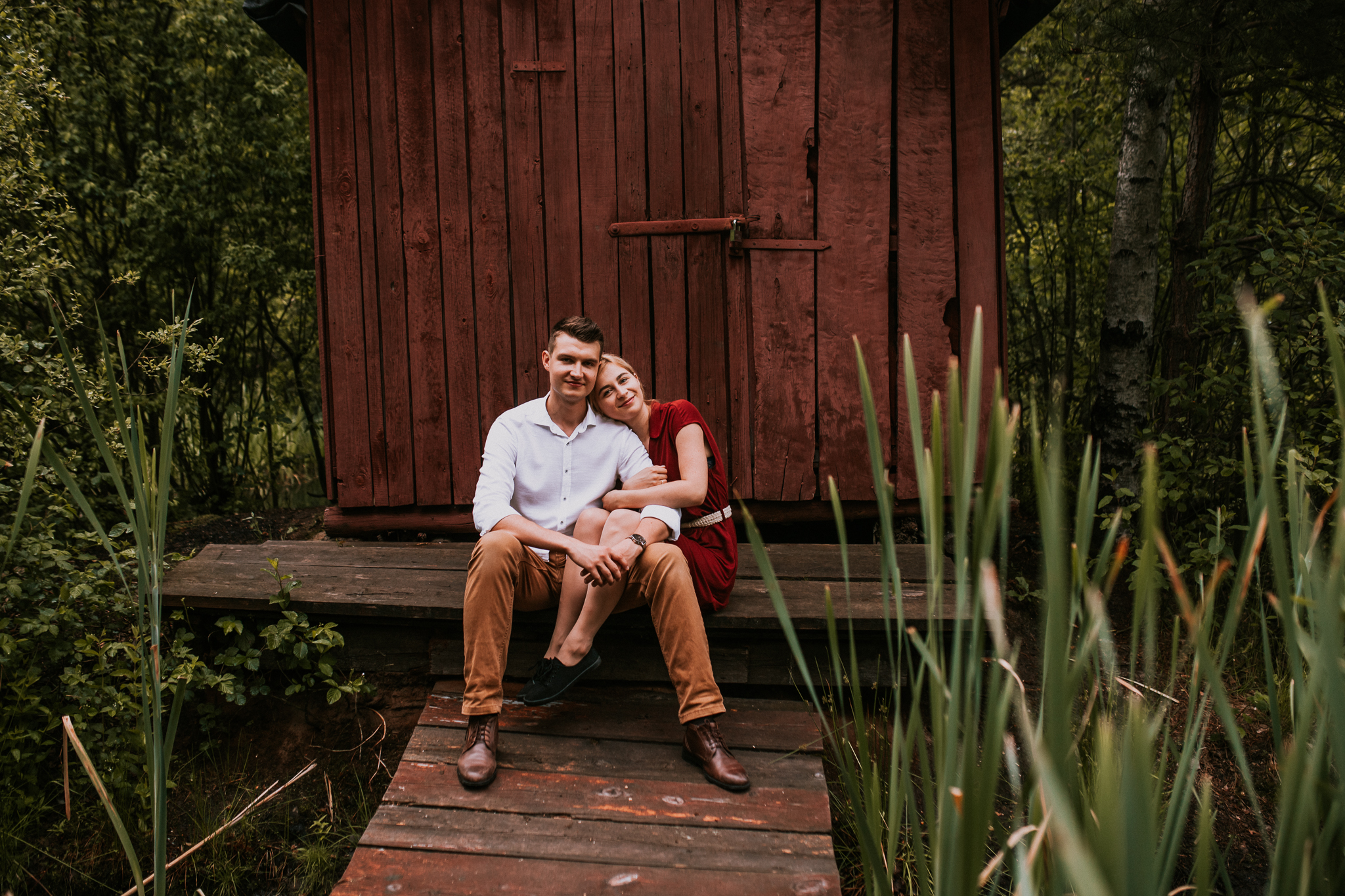 Kobieta i mężczyzna siedzą na wejściu do drewnianego domku. Dziewczyna ma nogi na kolanach mężczyzny, jest w niego wtulona