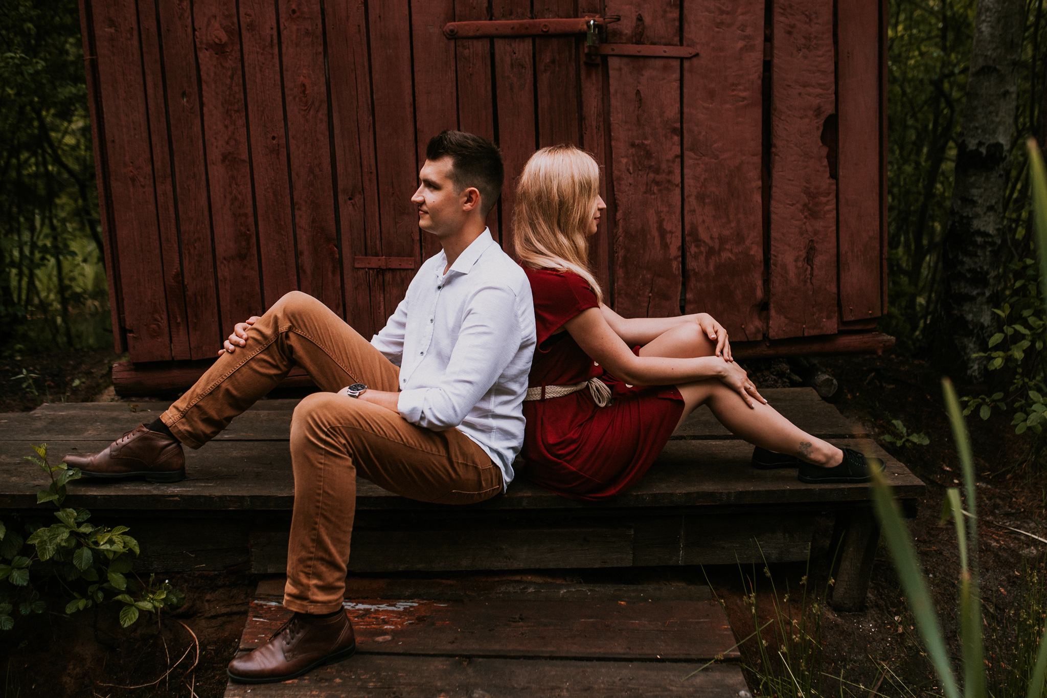 Mężczyzna i kobieta siedzą do siebie plecami. Są uśmiechnięci i patrzą przed siebie. Sesja narzeczeńska w plenerze