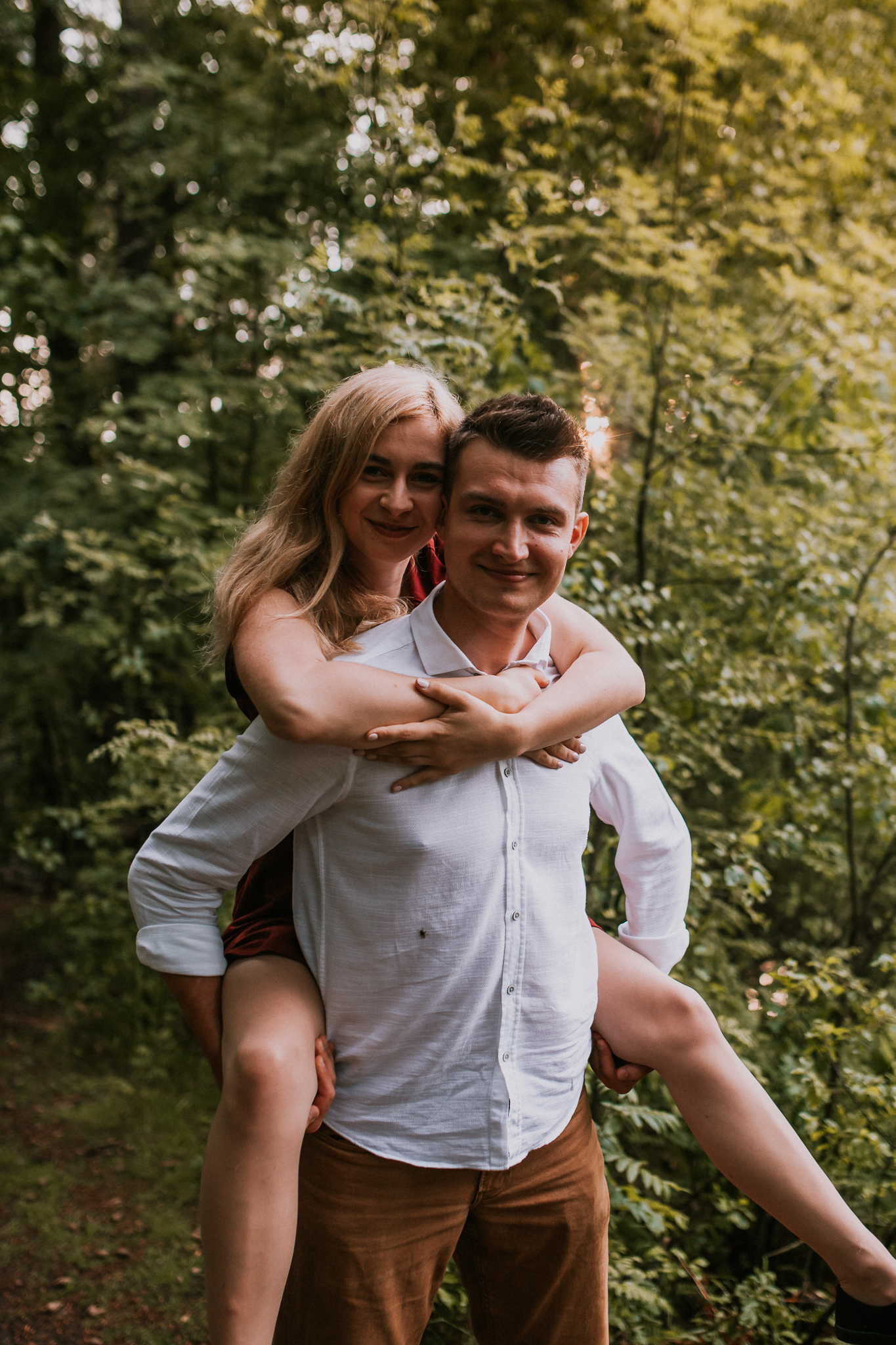 Chłopak niesie dziewczynę na plecach. W tle las. Sesja narzeczeńska w plenerze