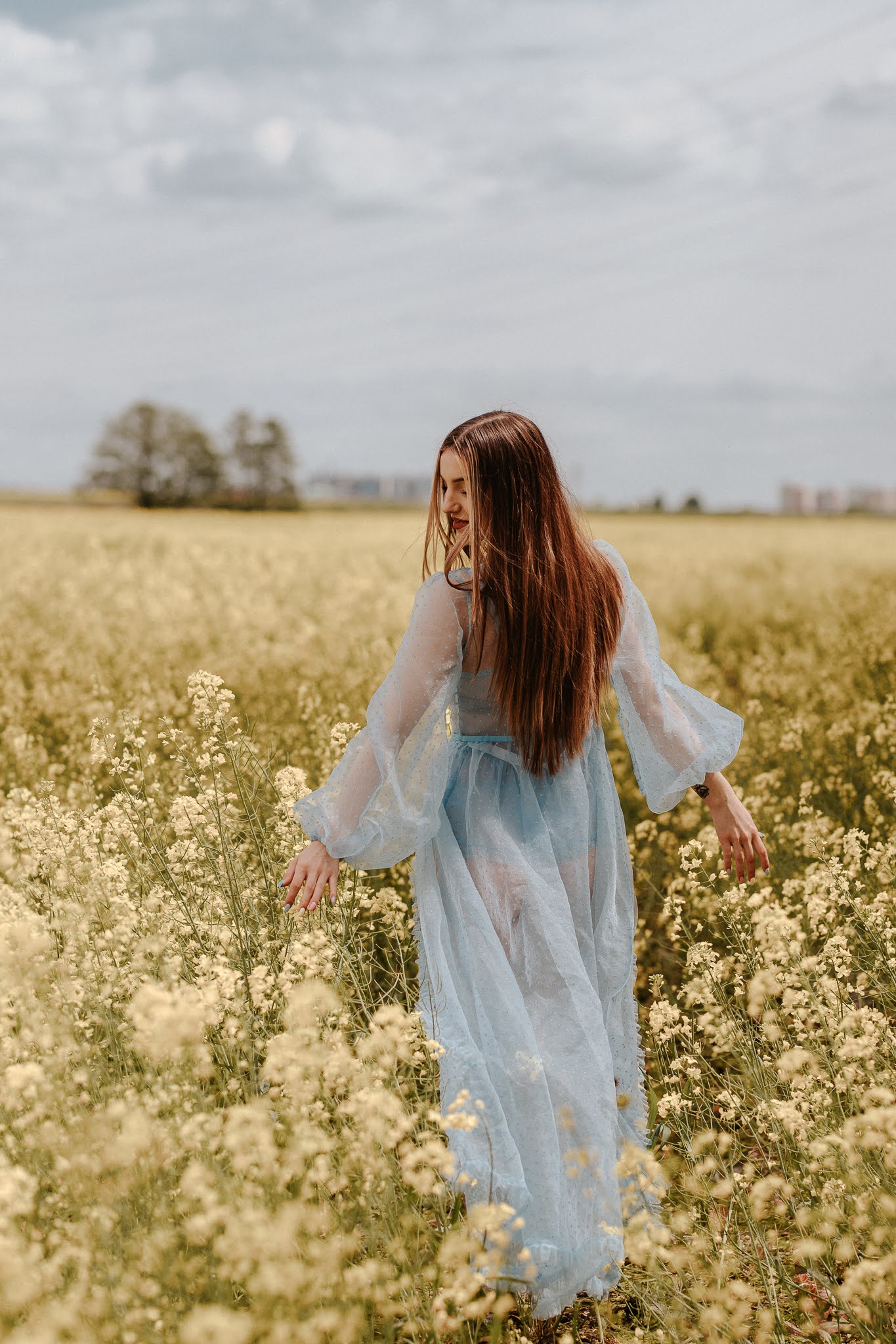 Dziewczyna stojąca tyłem w rzepaku w delikatnej muślinowej, błękitnej sukience