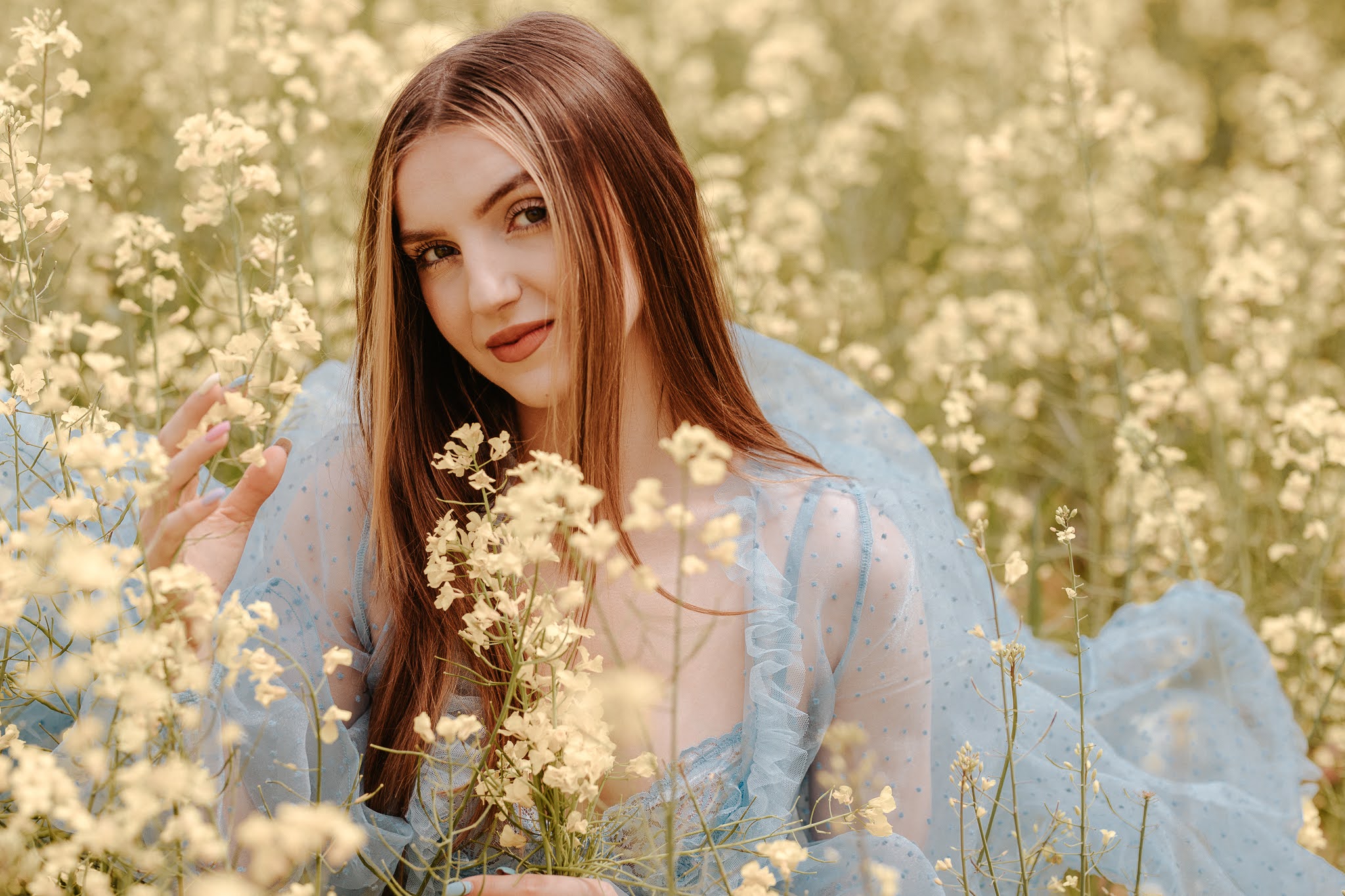 Uśmiechnięta dziewczyna siedząca w rzepaku, delikatnie spoglądająca w stronę obiektywu