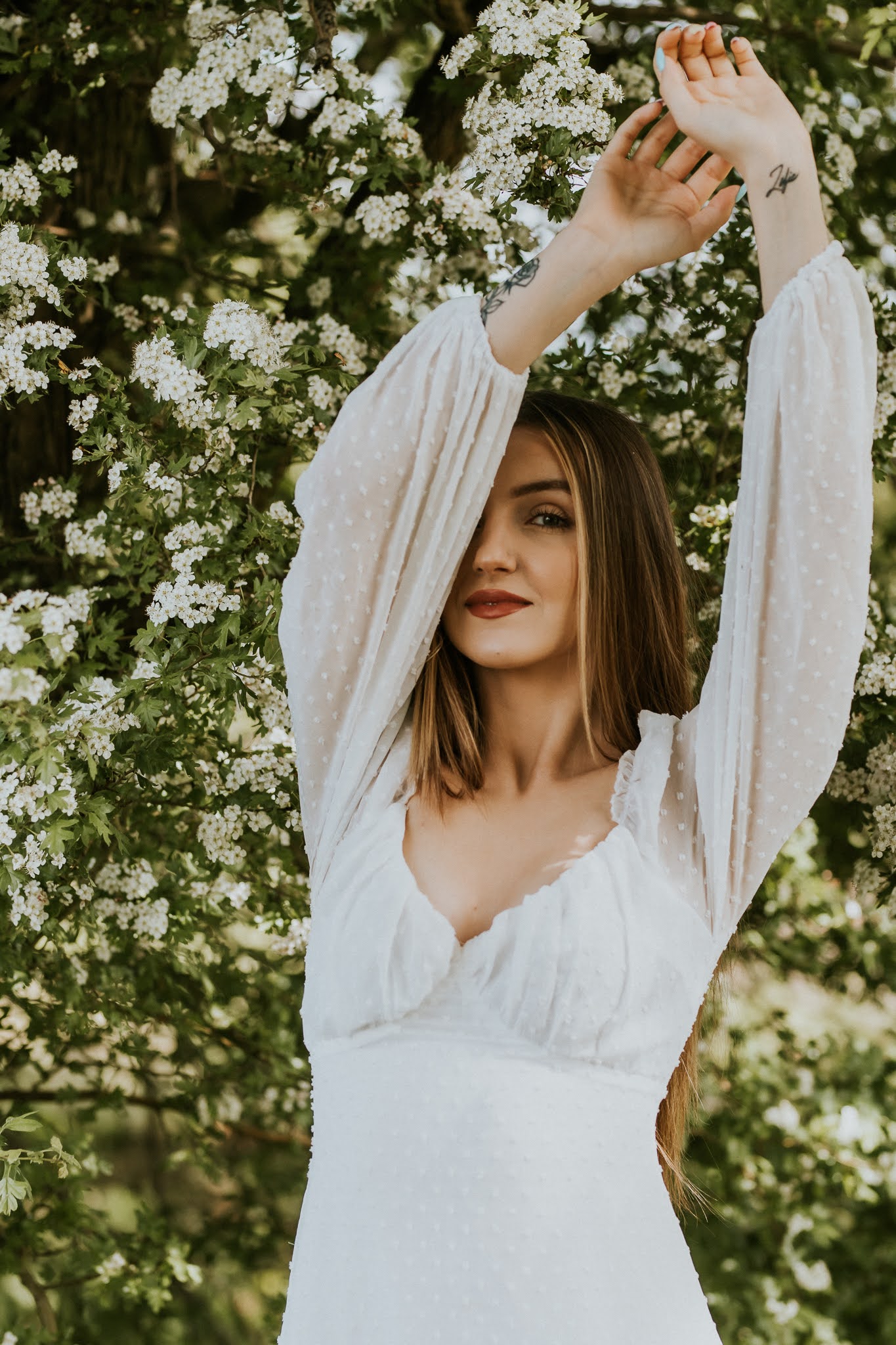 Dziewczyna z wyciągniętymi rękoma. Zasłania część swojej twarzy lewą ręką. Na tle kwitnącego drzewa