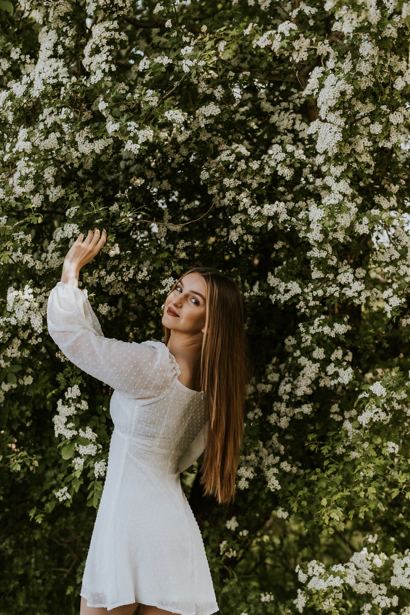 Dziewczyna w białej sukience jedną ręką opiera się o drzewo i patrzy w tył