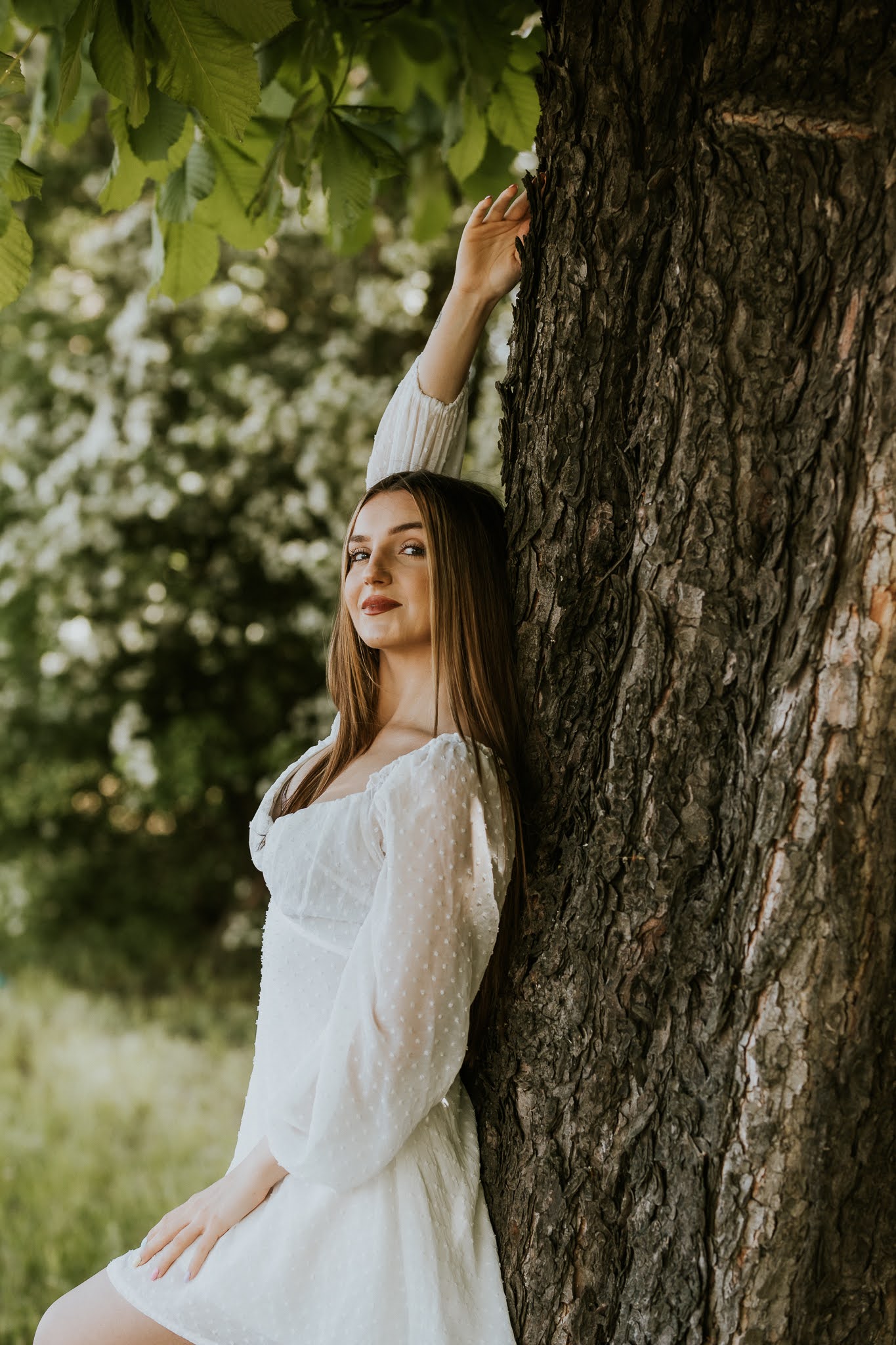 Dziewczyna w bieli oparta o drzewo z wyciągiętą prawą ręką