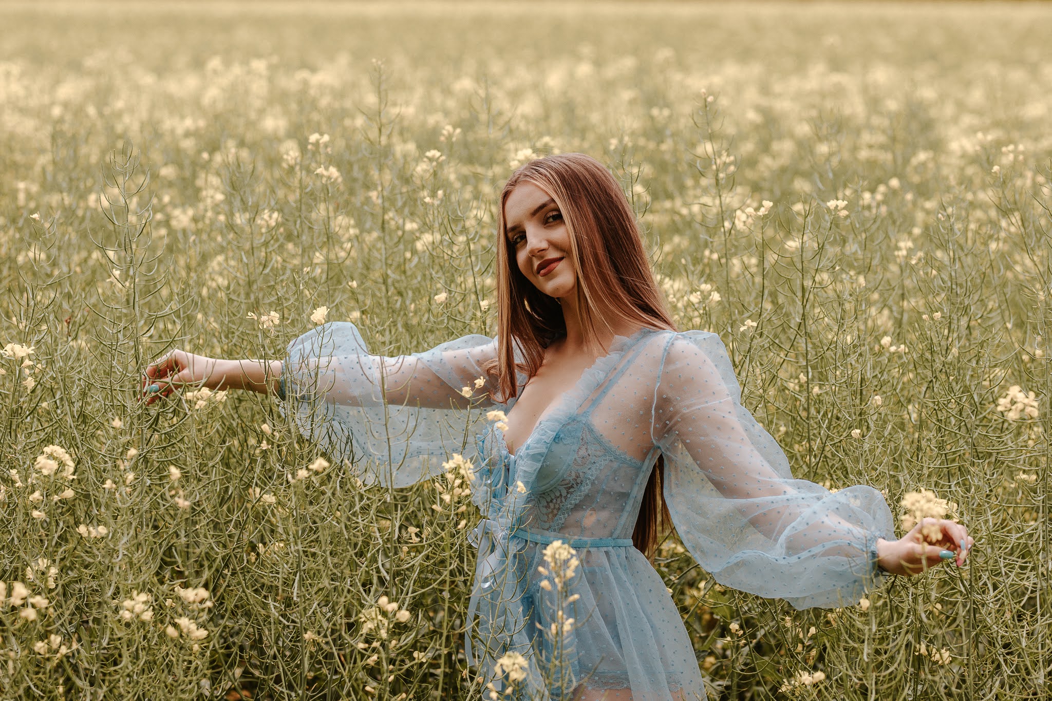 Młoda kobieta w błękitnej sukience stojąca w rzepaku z wyciągniętymi rękoma