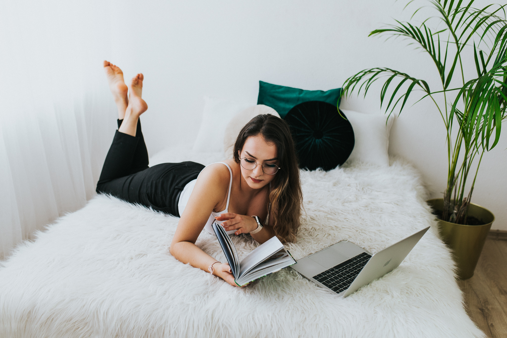 Kobieta leży na łóżku i przegląda książkę, obok stoi laptop.