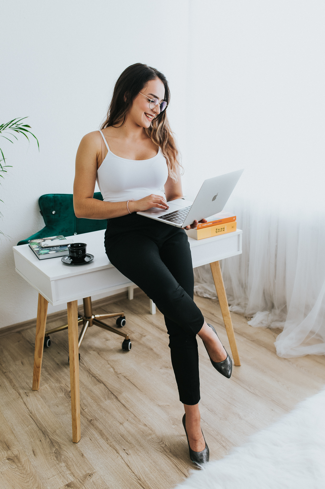 Usmiechnięta kobieta oparta bokiem o biurko. W rękach trzyma laptopa.