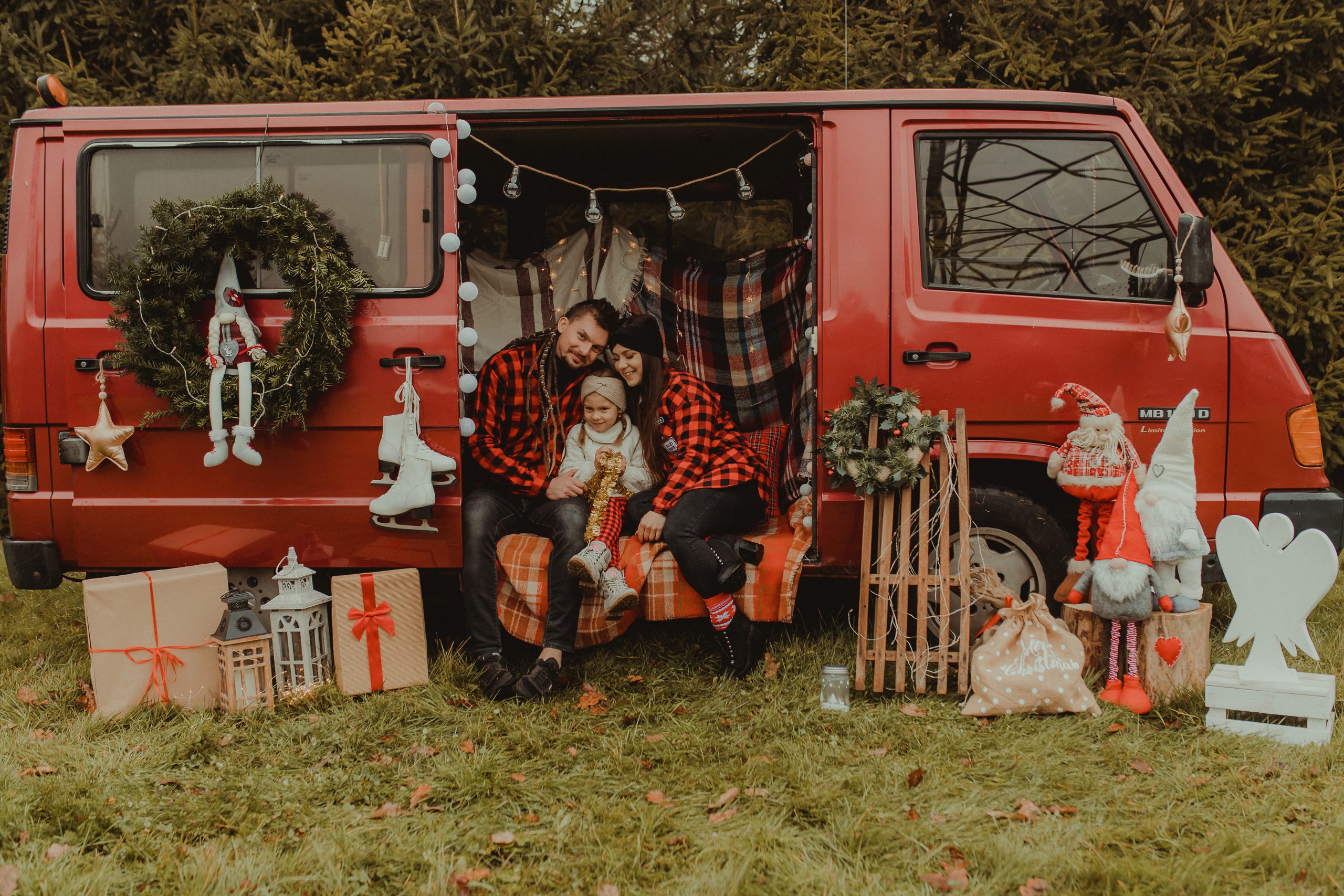 Sesja świąteczna. Czerwony, przystrojony świątecznie bus w środku dwoje dorosłych ludzi i mała dziewczynka, siedzą na kocu