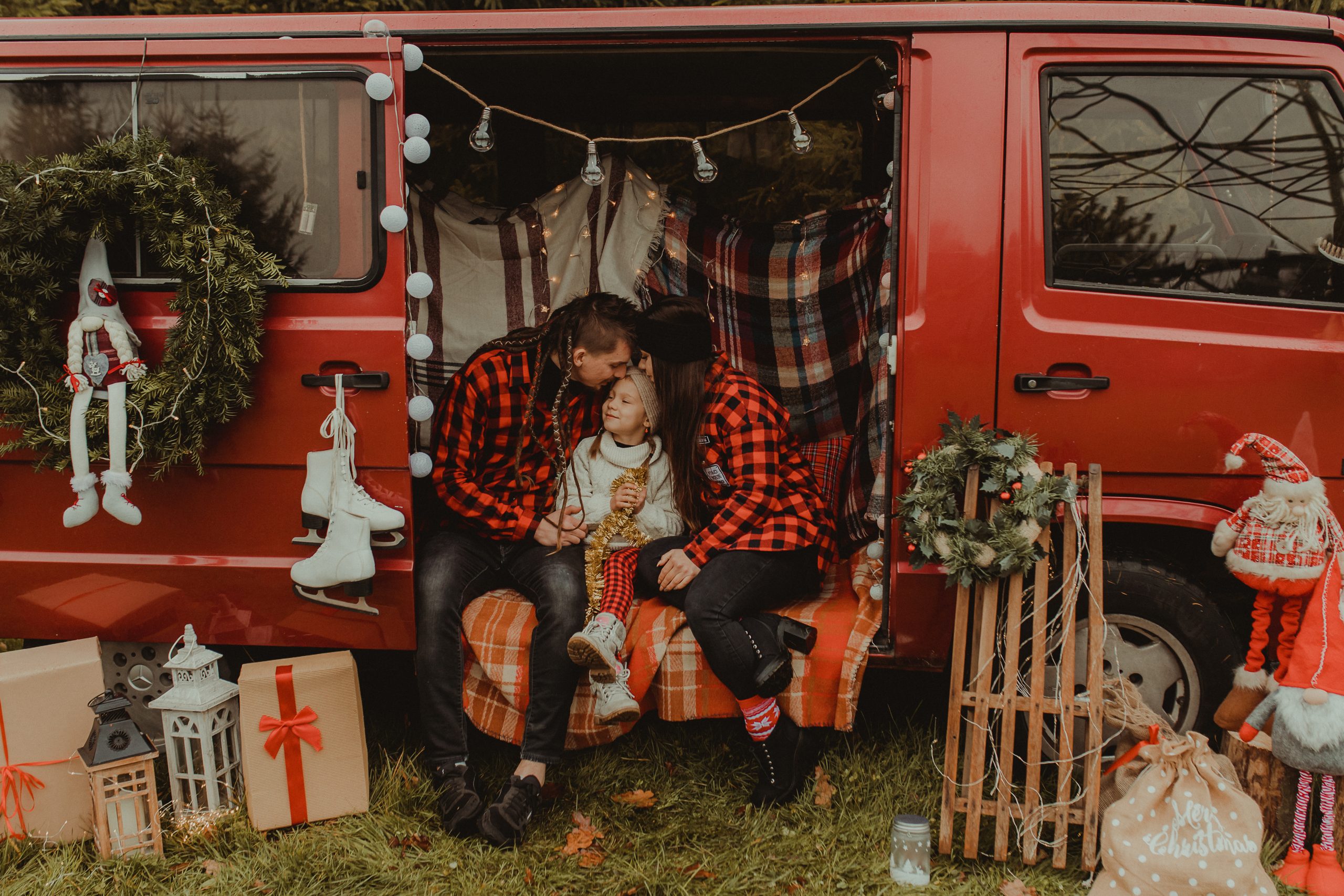 Czerwony bus wystrojony świątecznie. W środku siedzi rodzina. Rodzice całują dziecko w czoło. Sesja świąteczna