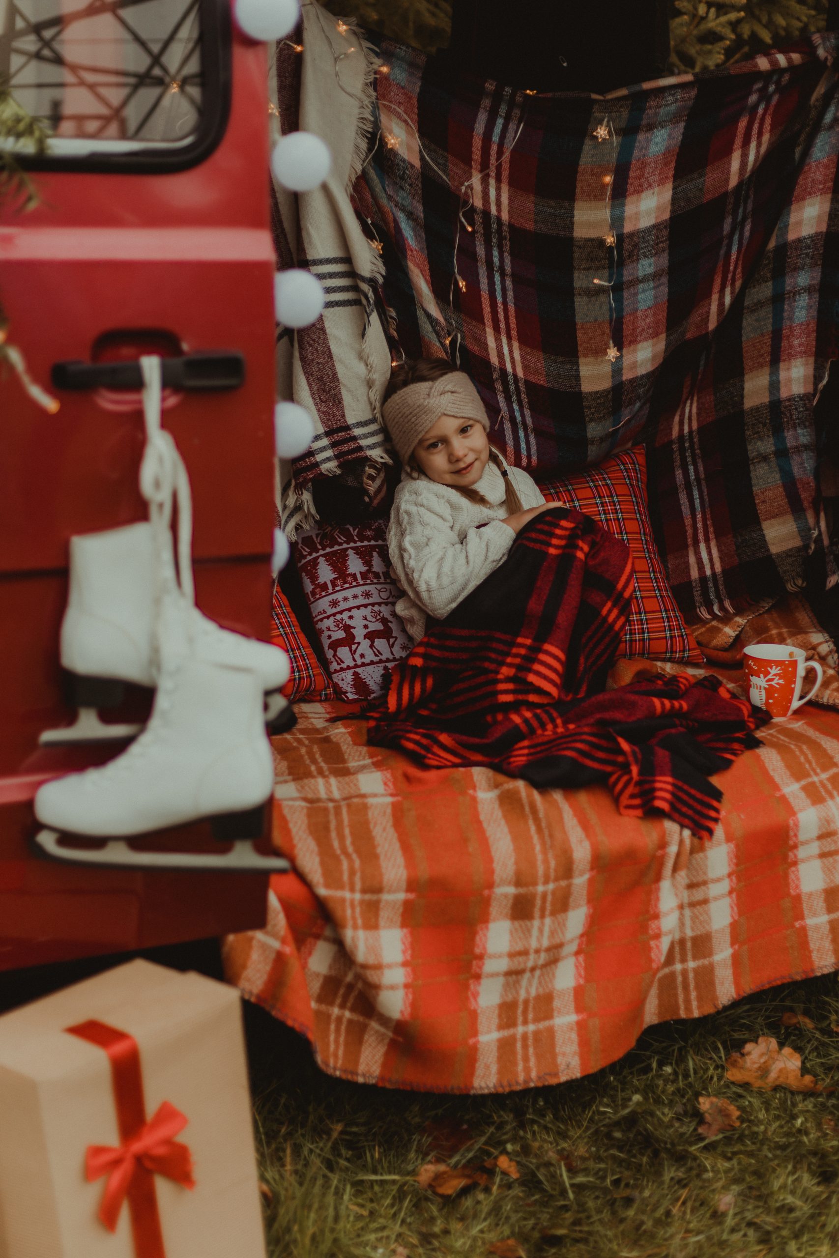 Sesja świąteczna. Dziewczynka siedzi w busie otulona czerwonym kocem. Na klamce samochodu wiszą białe łyżwy