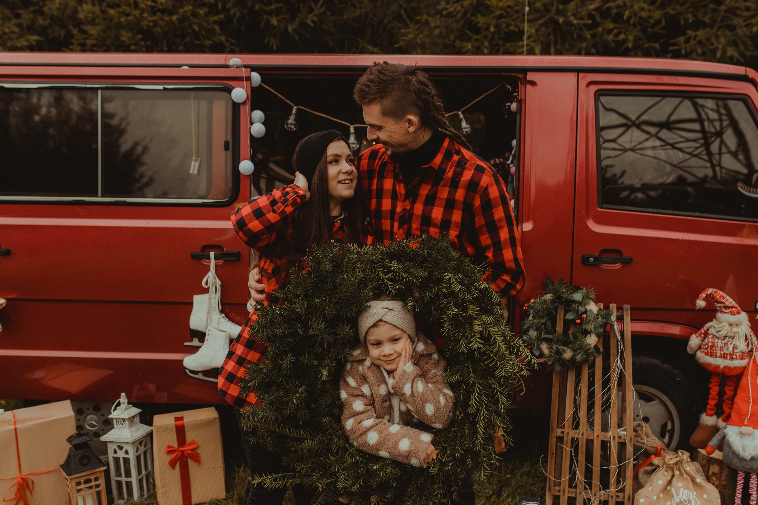 Mała dziewczynka stoi we wieńcu, który trzymają rodzice ubrani w czerwone kraciaste koszule. Sesja świąteczna