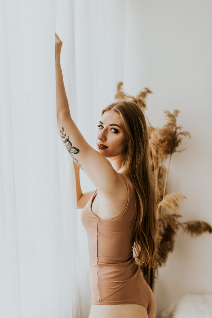 kobieta w body stoi przy oknie i patrzy się w stronę obiektywu