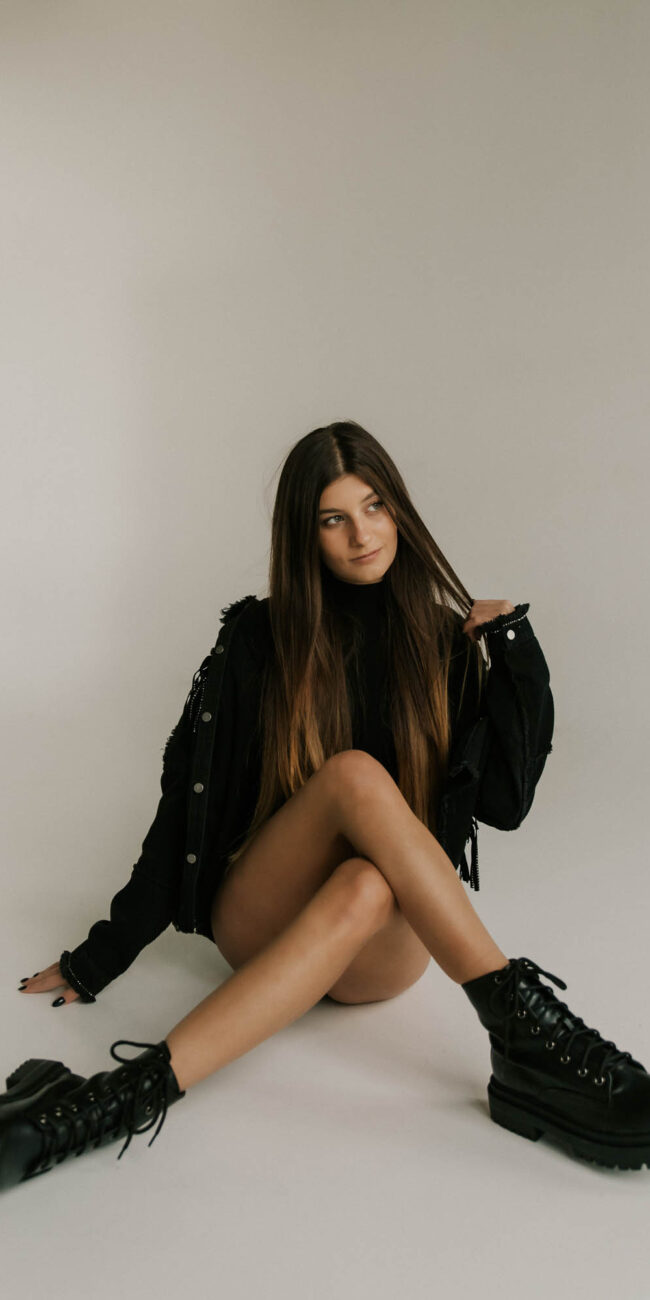 Brunetka siedząca przy ścianie z założonymi nogami w czarnej stylizacji, ma gołe nogi, Fusialka