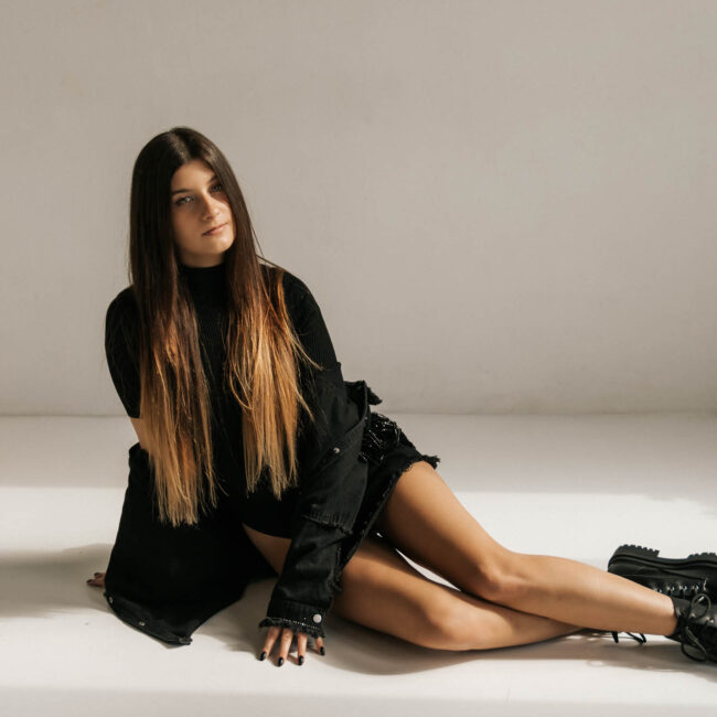 Kobieta w czarnej stylizacji siedzi na podłodze i patrzy w obiektyw, ma długie włosy