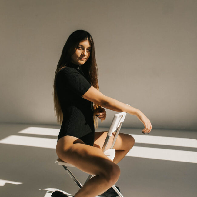 Dziewczyna siedząca na krześle . Ma czarne body i gołe nogi oraz ciężkie buty.