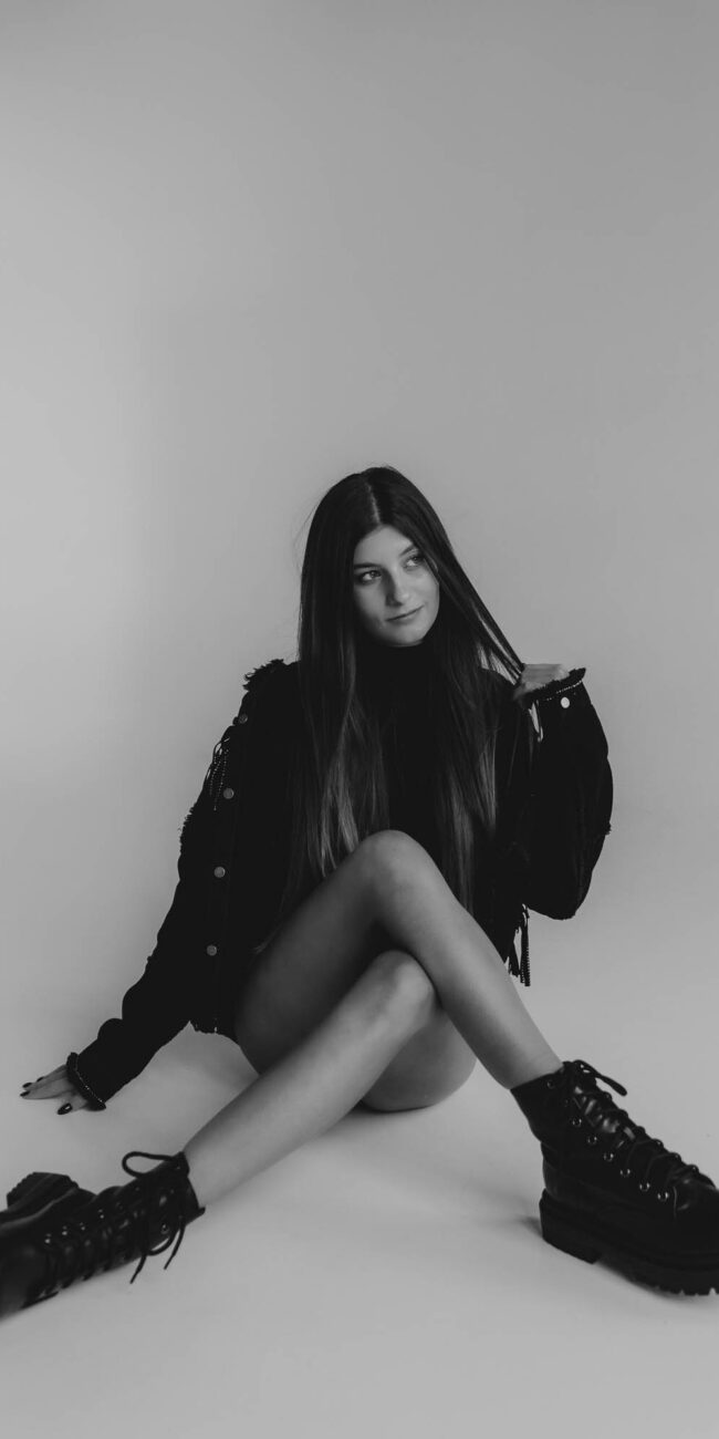 Brunetka siedząca przy ścianie z założonymi nogami w czarnej stylizacji, ma gołe nogi, Fusialka, fotografia czarno- biała