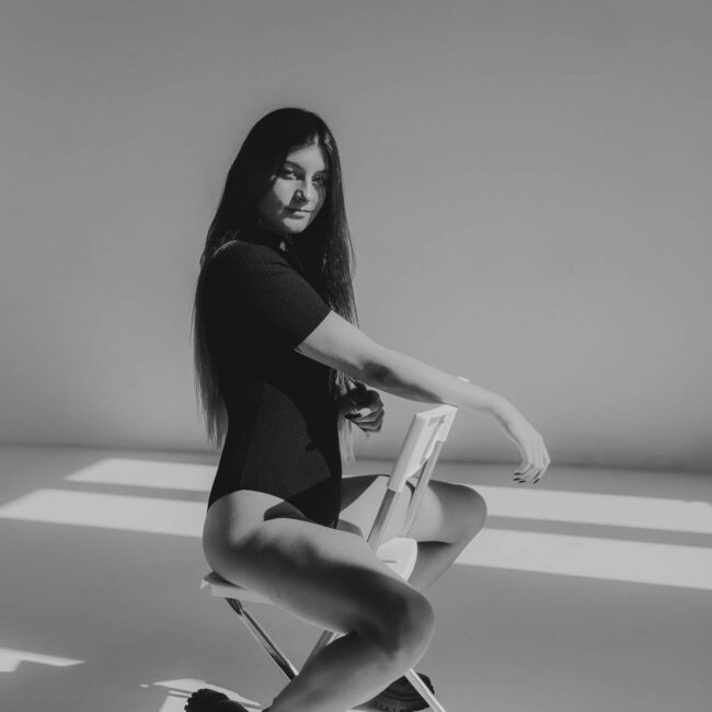 Dziewczyna siedząca na krześle . Ma czarne body i gołe nogi oraz ciężkie buty. Zdjęcie czarno- białe.