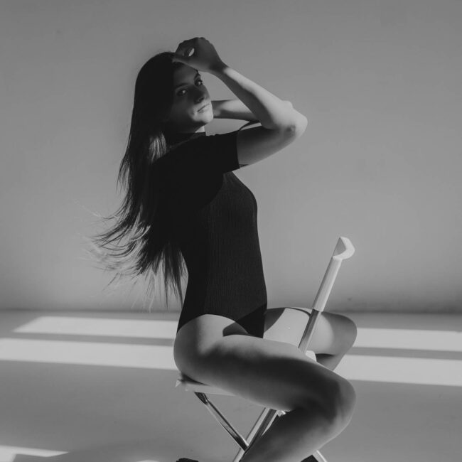 Dziewczyna w body siedzi na białym krześle i rękę trzyma na skroni, patrzy w obiektyw. Zdjęcie czarno- białe