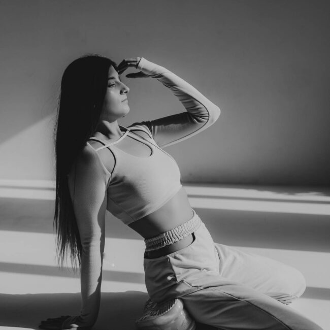 Brunetka w beżowym dresie siedzi na podłodze i trzyma rękę na skroni, drugą podpiera się o podłogę. Zdjęcie czarno- białe.