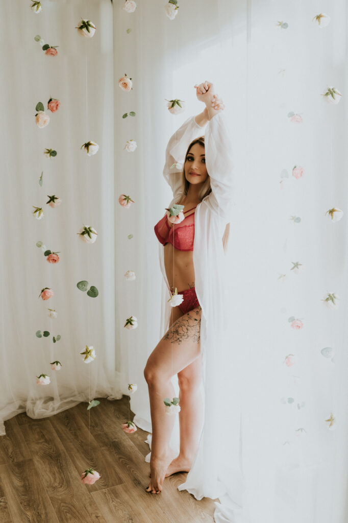 kobieta stoi przy firanie jest ubrana w bieliznę i narzutkę z góry zwisają kwiaty