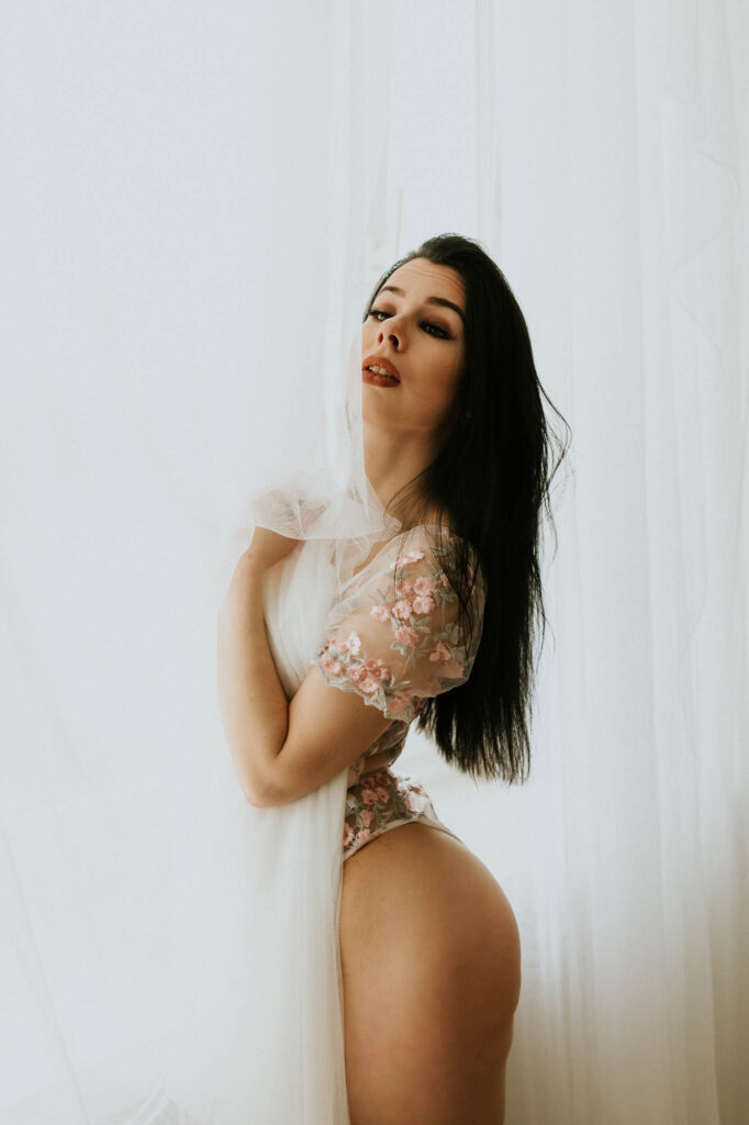 Kobieta w koronkowym białym body stoi przy firanie