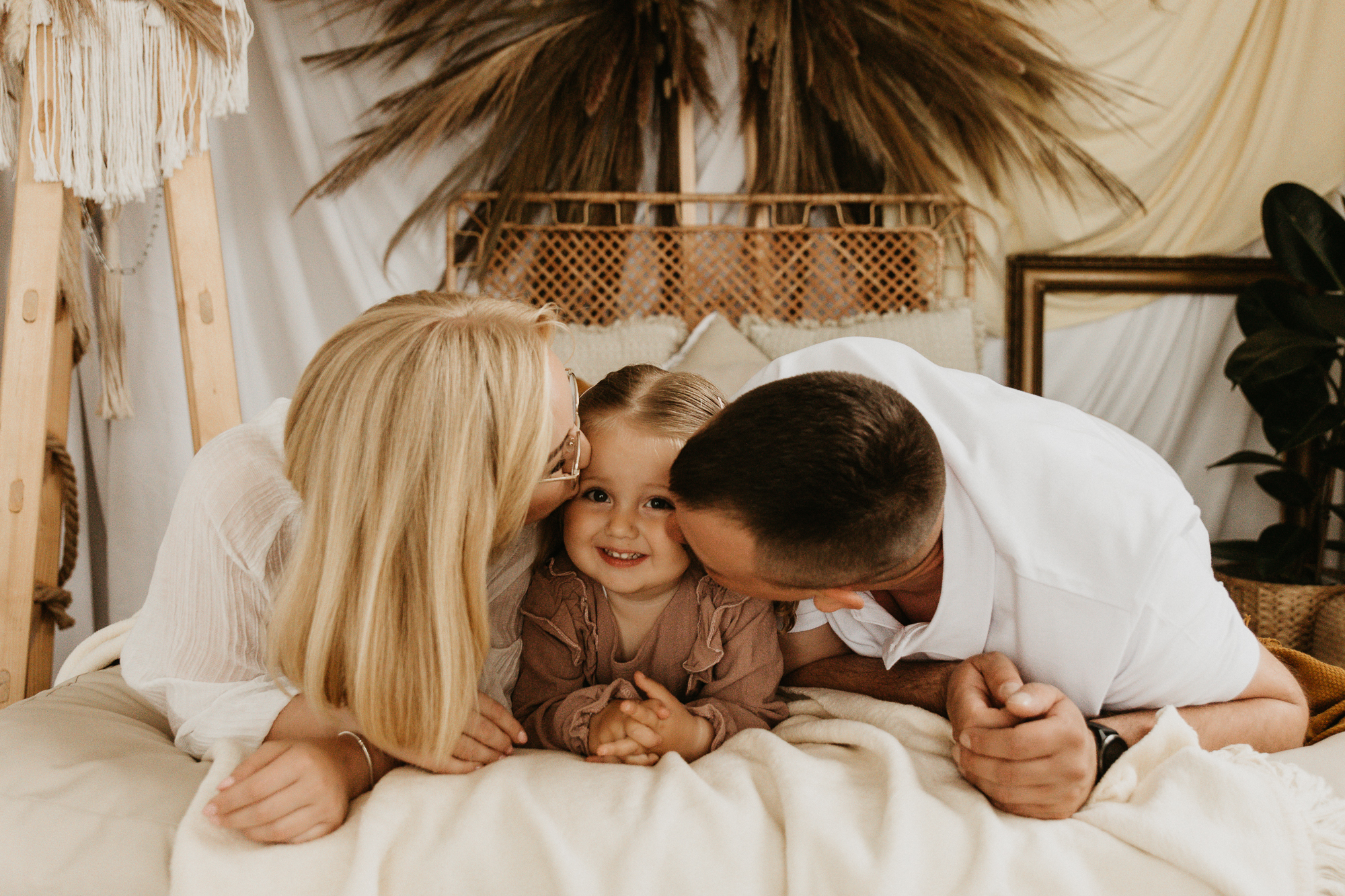 mężczyzna i kobieta całują dziewczynkę w policzek, leżą na łóżku, sesja rodzinna