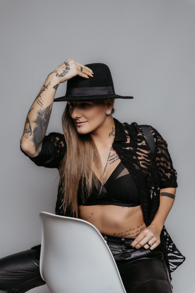 kobieta siedzi na krześle  ma czarny kapelusz i trzyma się wytatuwoaną ręką